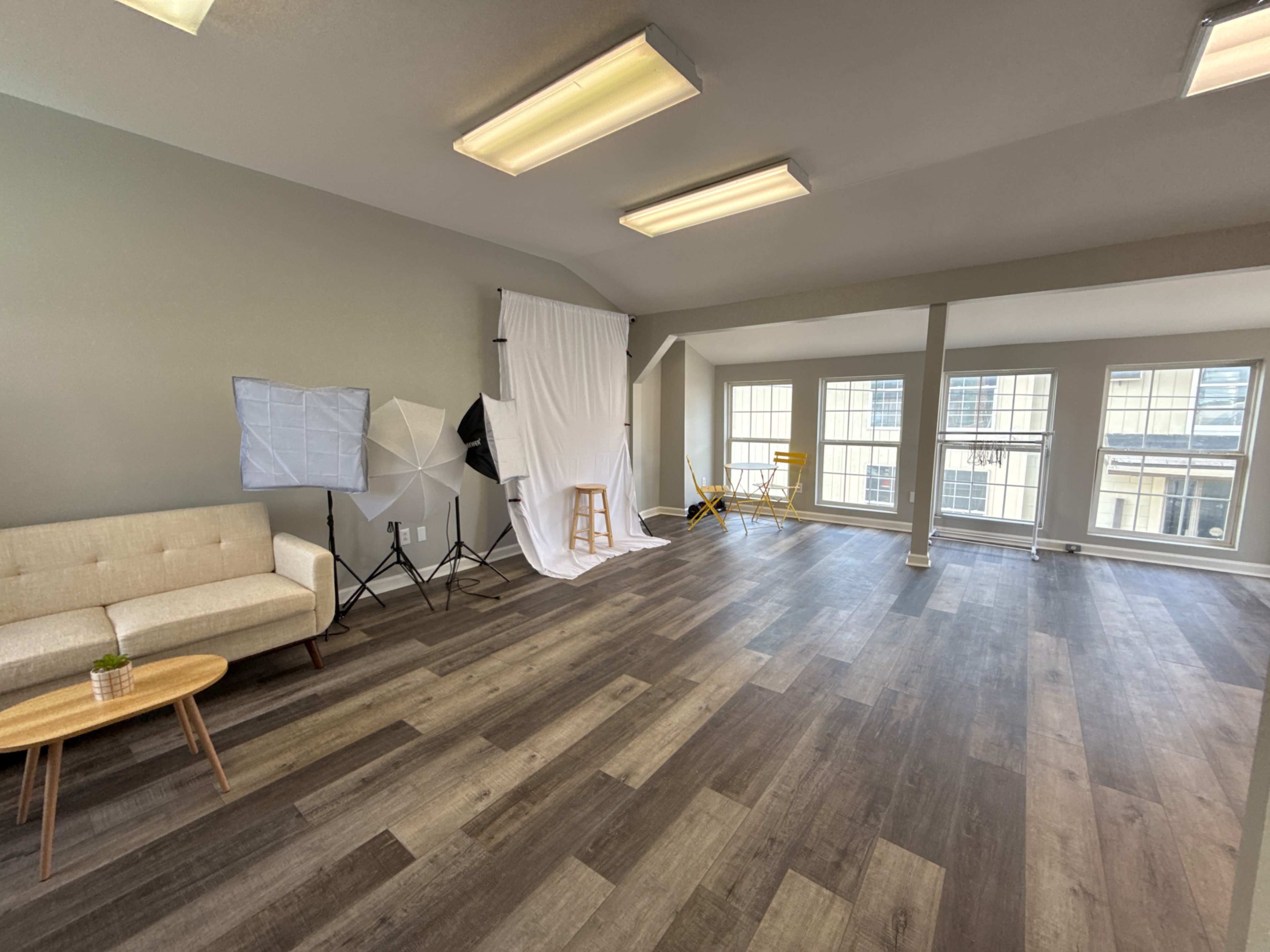 The image shows a spacious room with wooden flooring, featuring a couch, lighting equipment, and a white backdrop set up for photography.