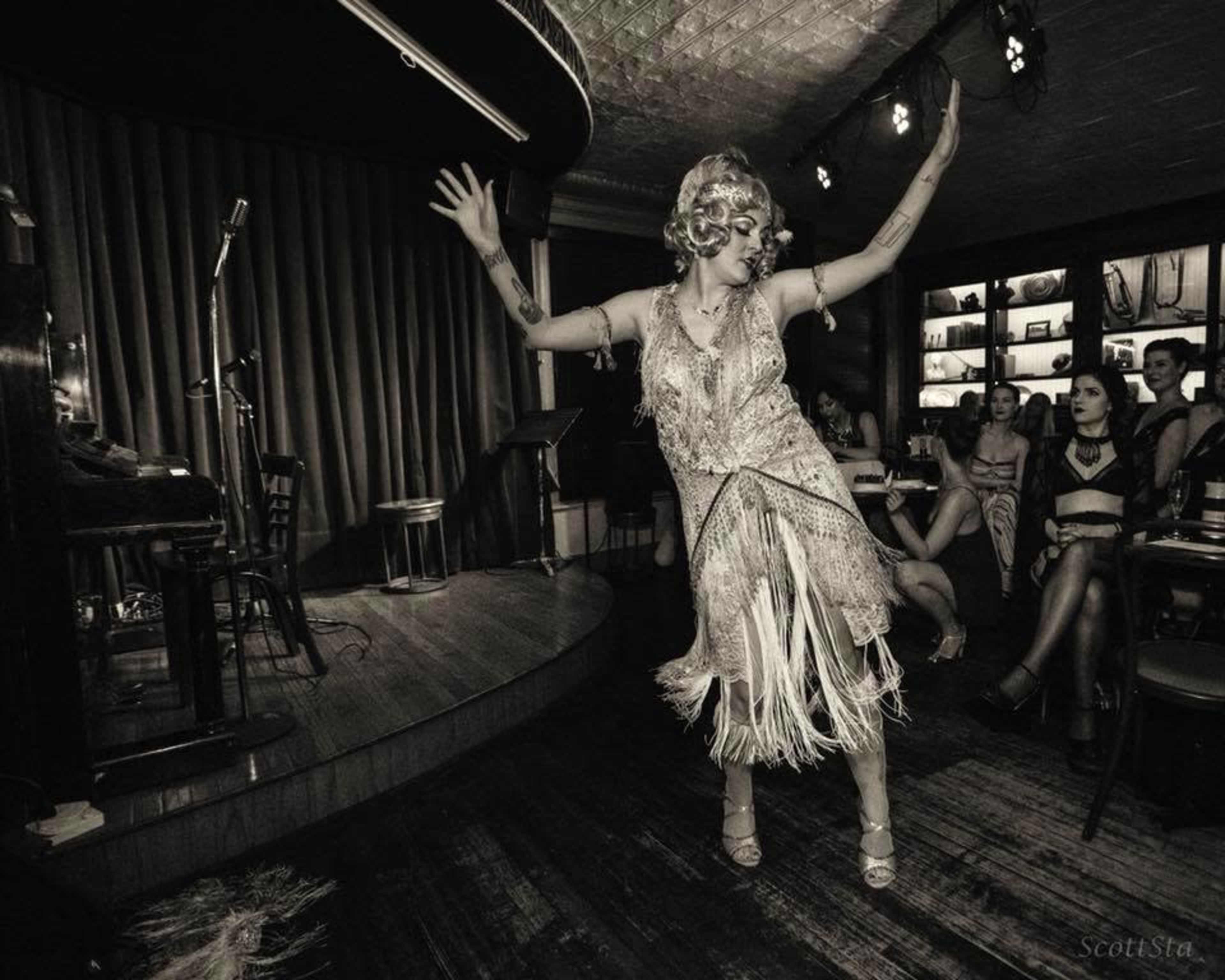 A performer in a fringed costume dances on stage in a cabaret setting, while an audience watches from their seats.