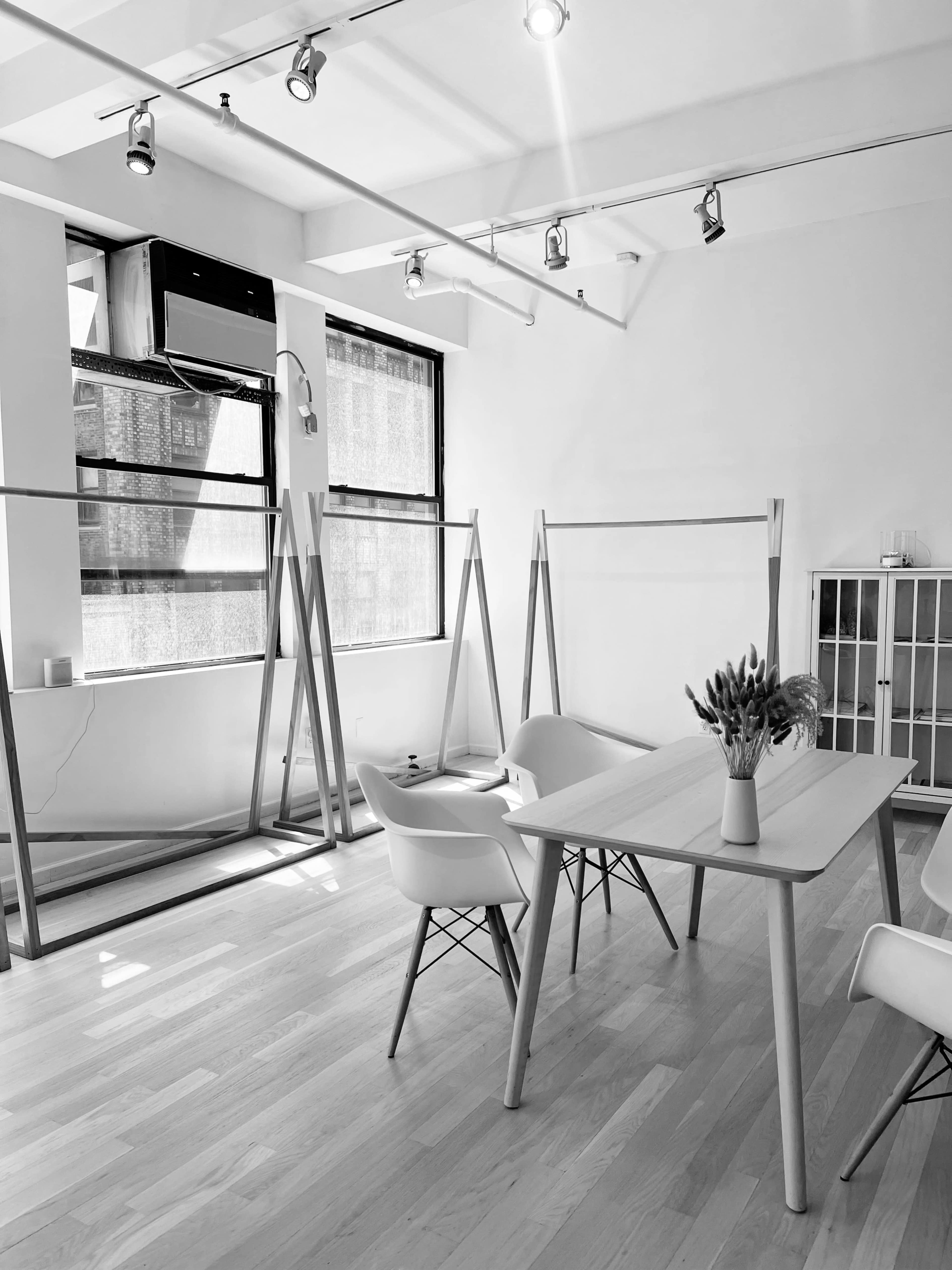 A minimalist interior space with two large windows, several clothing racks, a table with a vase of flowers, and a small cabinet.
