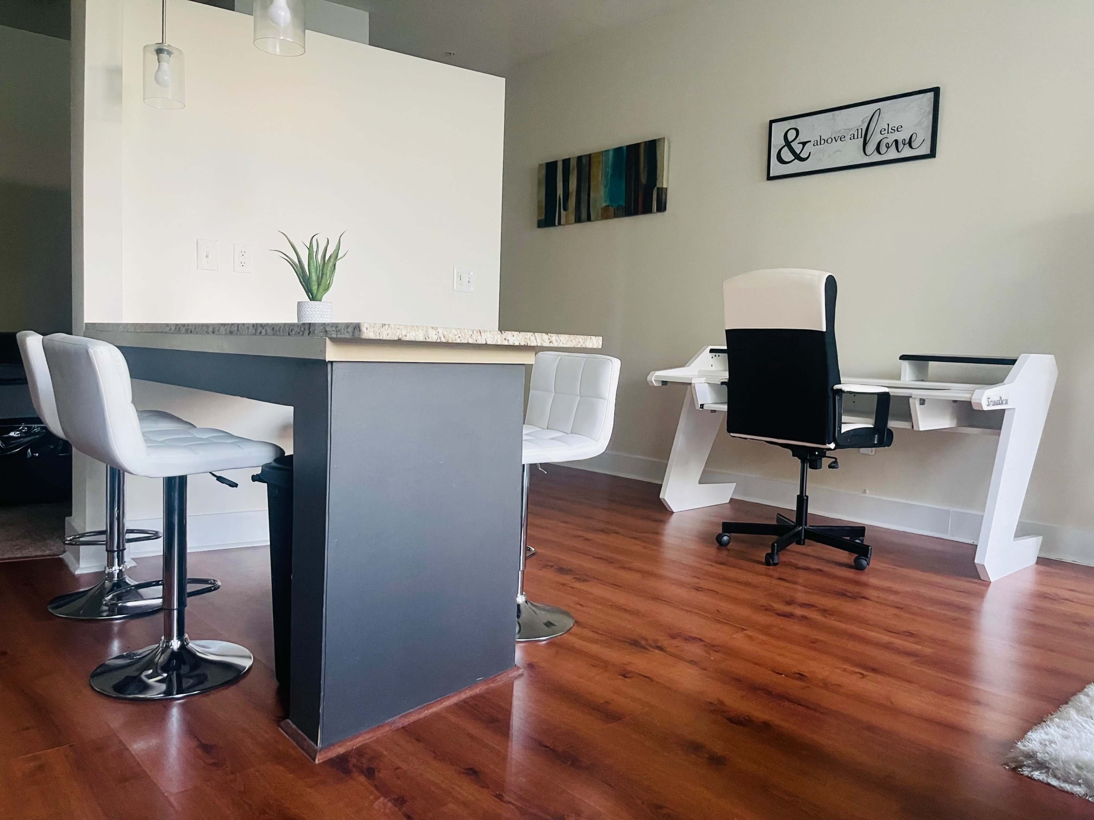 A modern interior featuring a small dining area with a granite-top table and four white stools, along with a workspace that includes a white desk and an office chair.