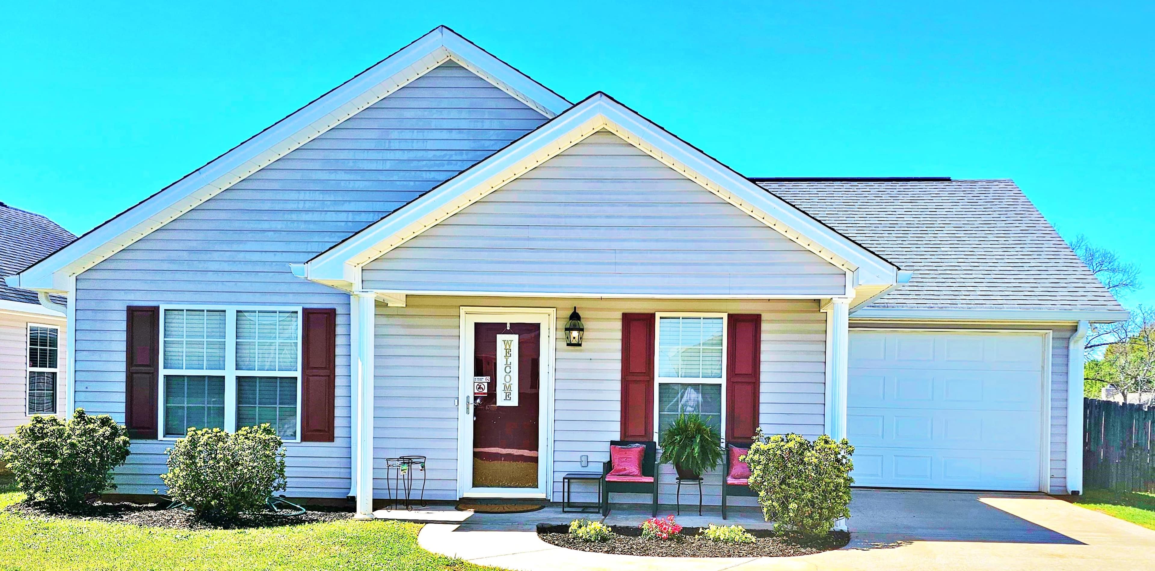 The image shows a modern single-story house with a peaked roof, large windows, and a well-maintained front yard featuring shrubs and decorative plants.