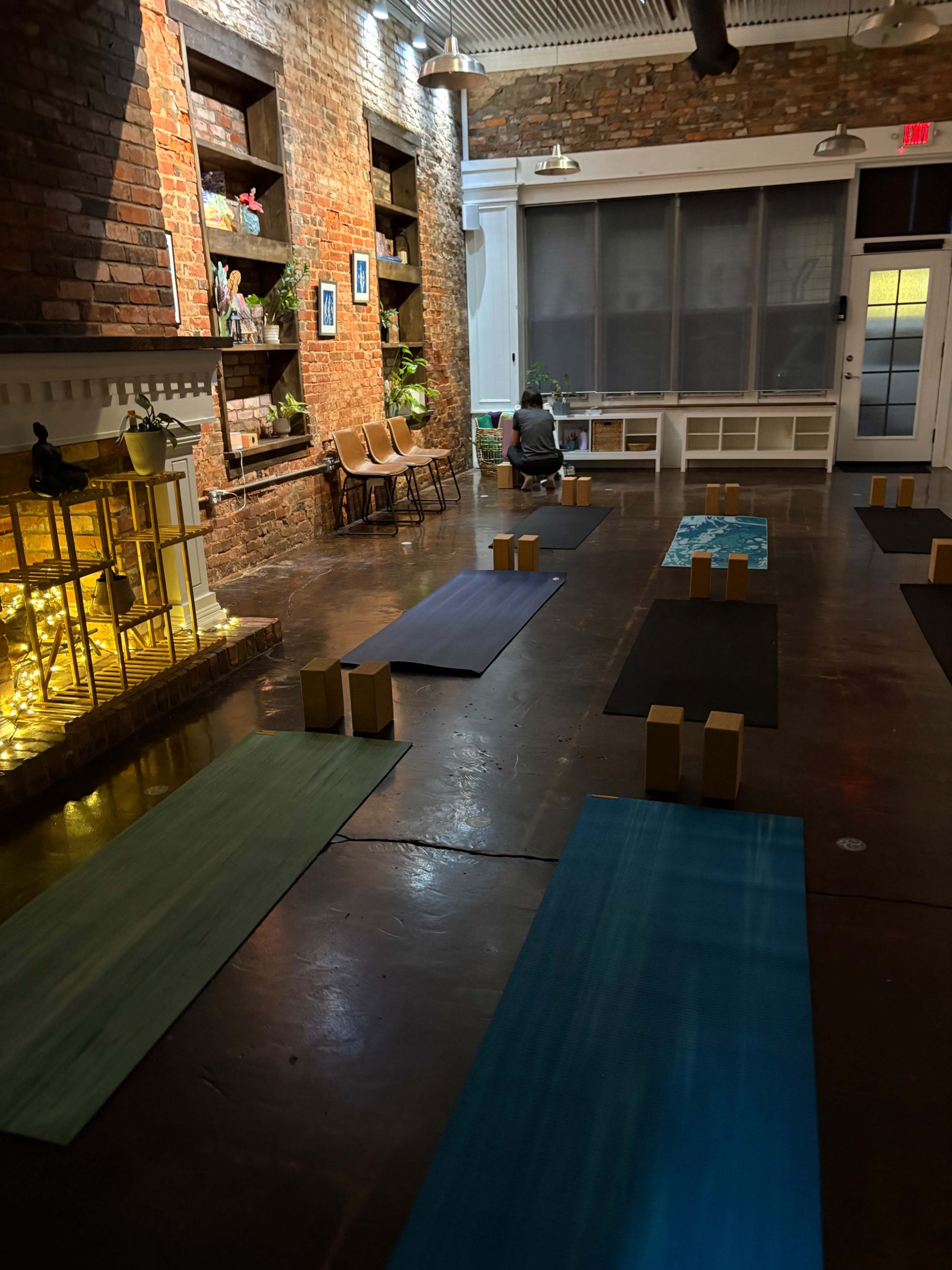 The image shows a yoga studio with several yoga mats arranged on a polished floor, wooden blocks positioned on the mats, and warm lighting highlighting exposed brick walls.