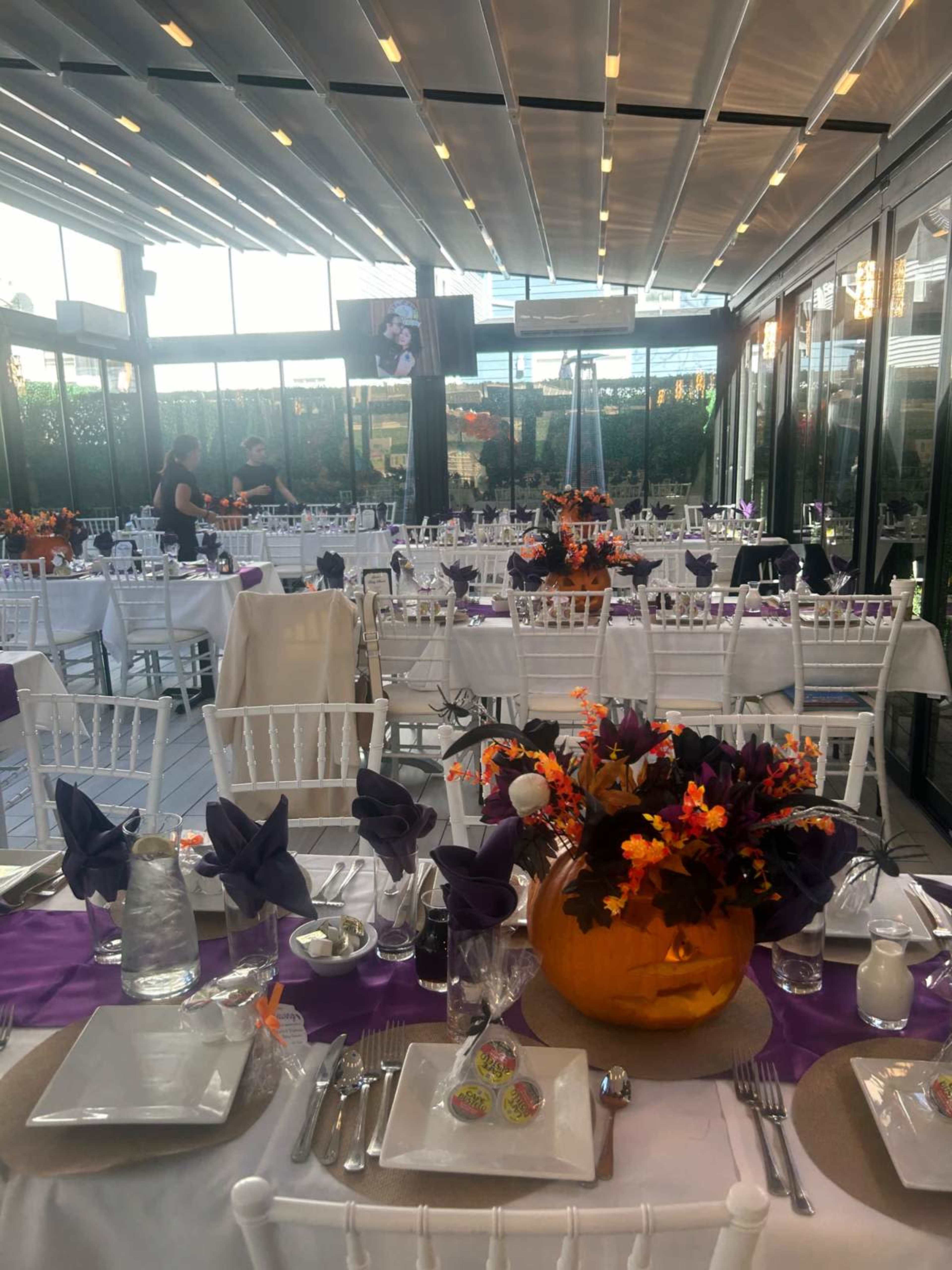 A decorated dining area features white chairs and tables set with purple and orange accents, complete with pumpkins and floral centerpieces.