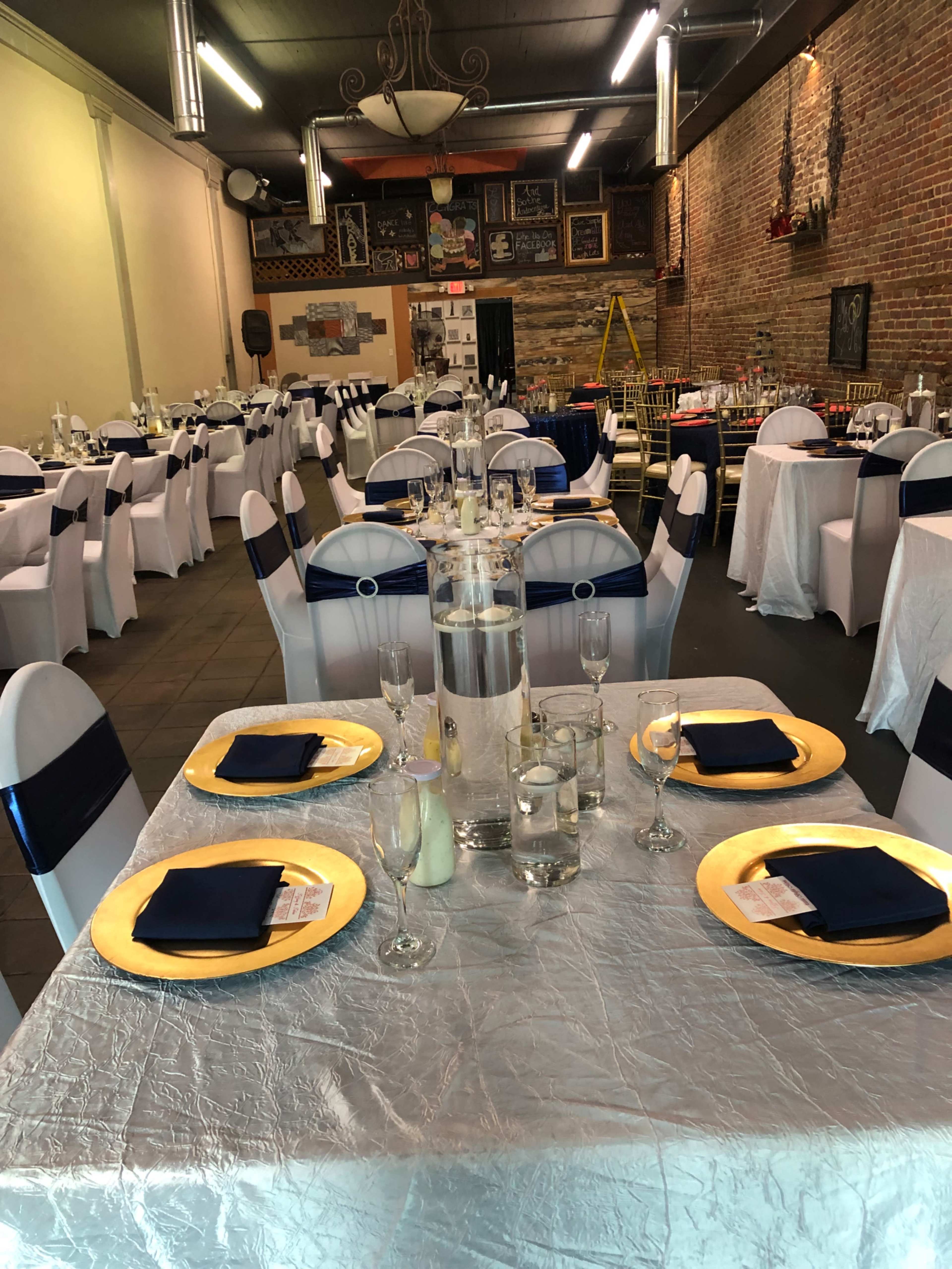 A banquet hall set for an event, featuring elegantly arranged tables with white tablecloths, gold chargers, and navy blue napkins, along with a brick wall and decorative elements in the background.