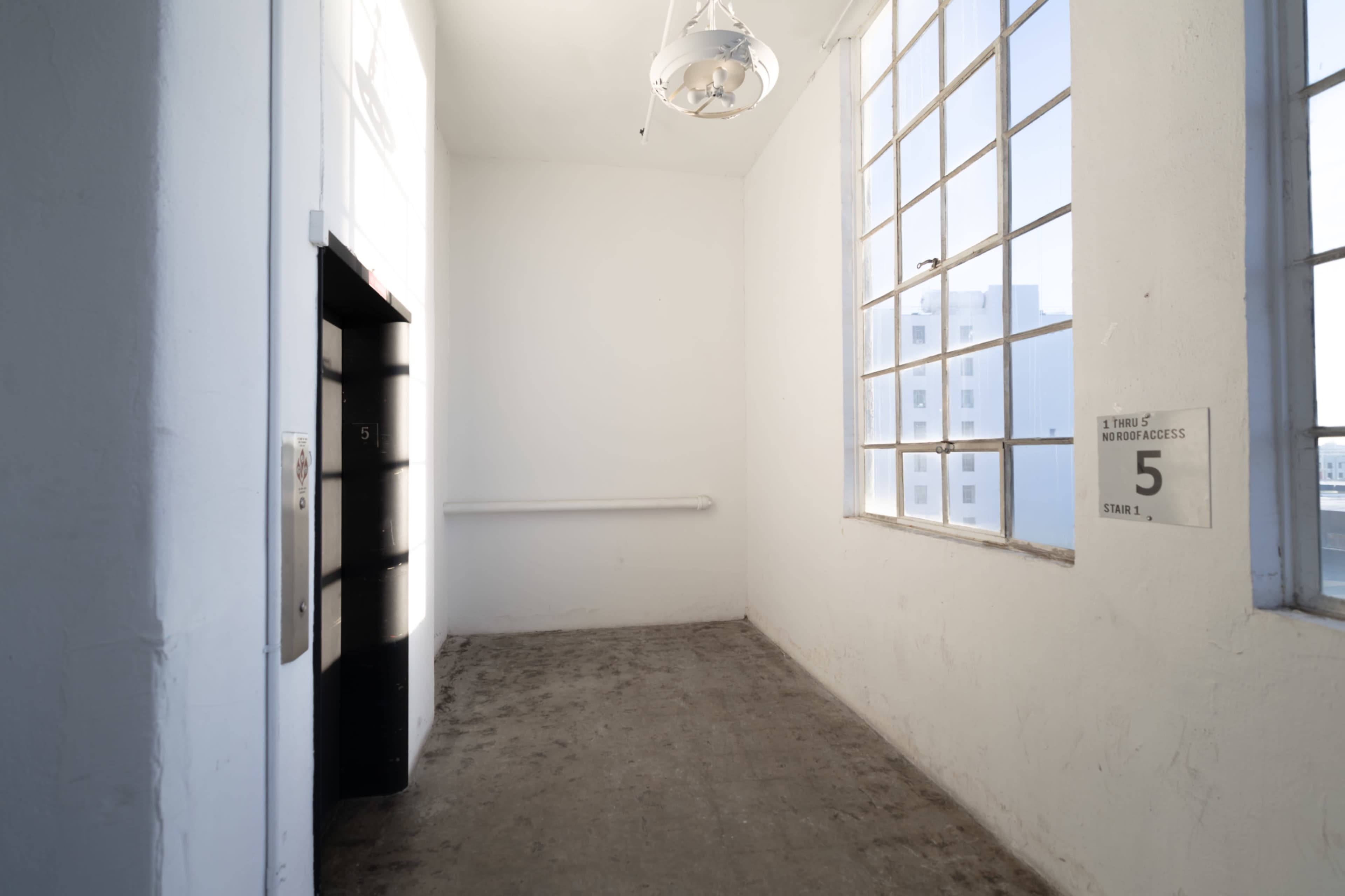 The image shows a spacious, empty room with large windows and a metal elevator door on one wall.