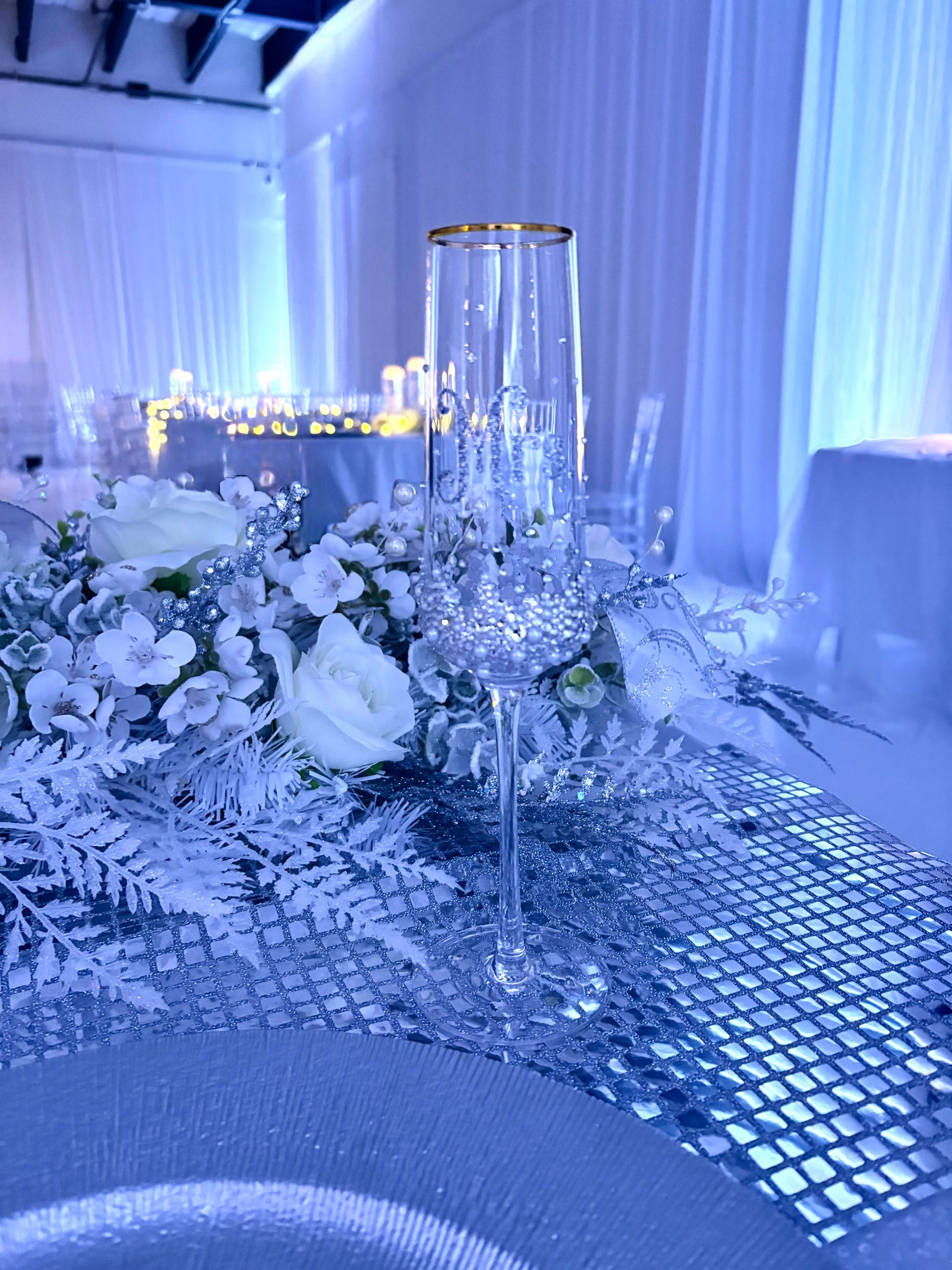 A decorated champagne flute stands on a shimmering table setting adorned with white roses and silver foliage under ambient blue lighting.