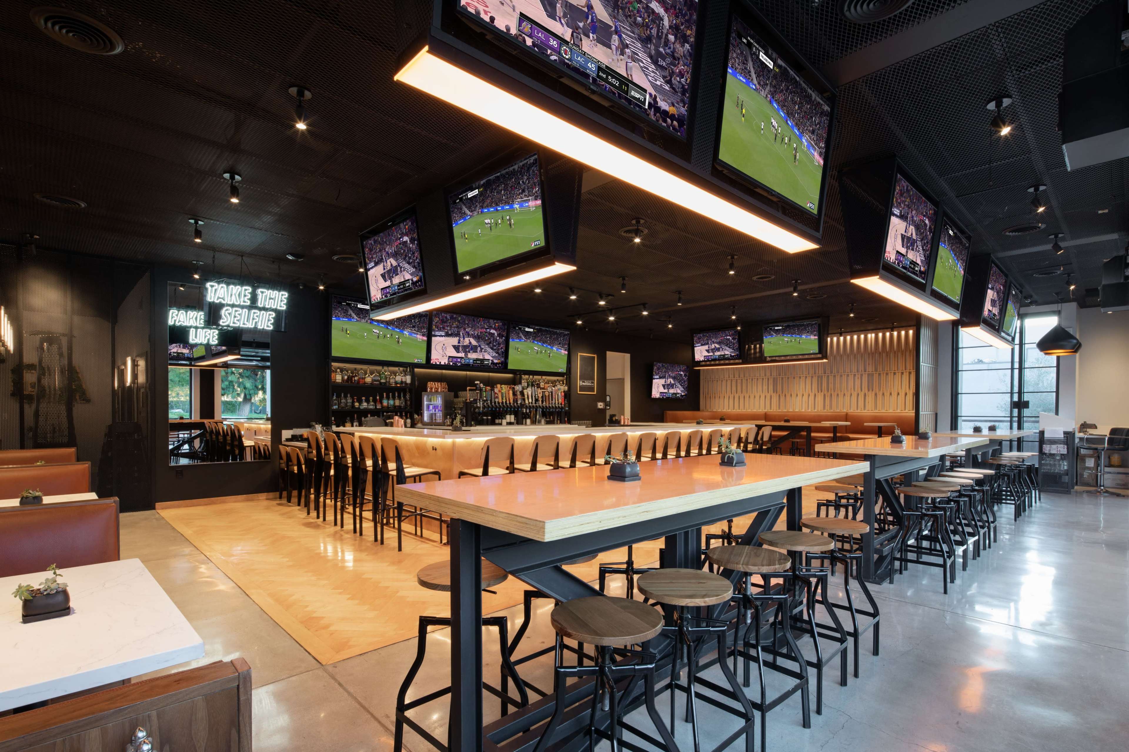 The image depicts a modern sports bar with multiple large screens displaying a basketball game, featuring a long bar counter with high stools and sleek wooden accents.