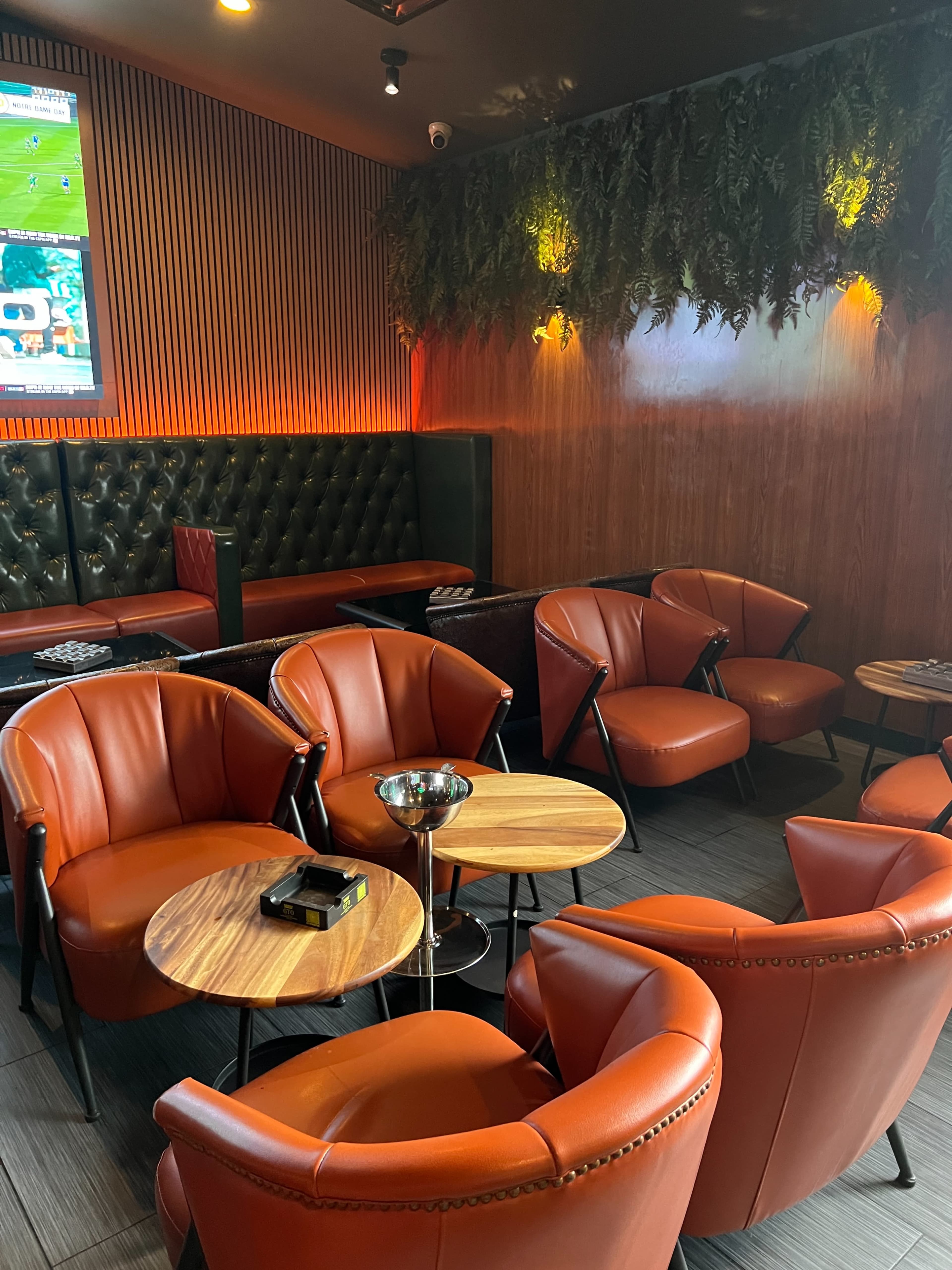 The image shows a cozy seating area in a lounge with orange chairs surrounding round tables, accented by wood paneling and ambient lighting.