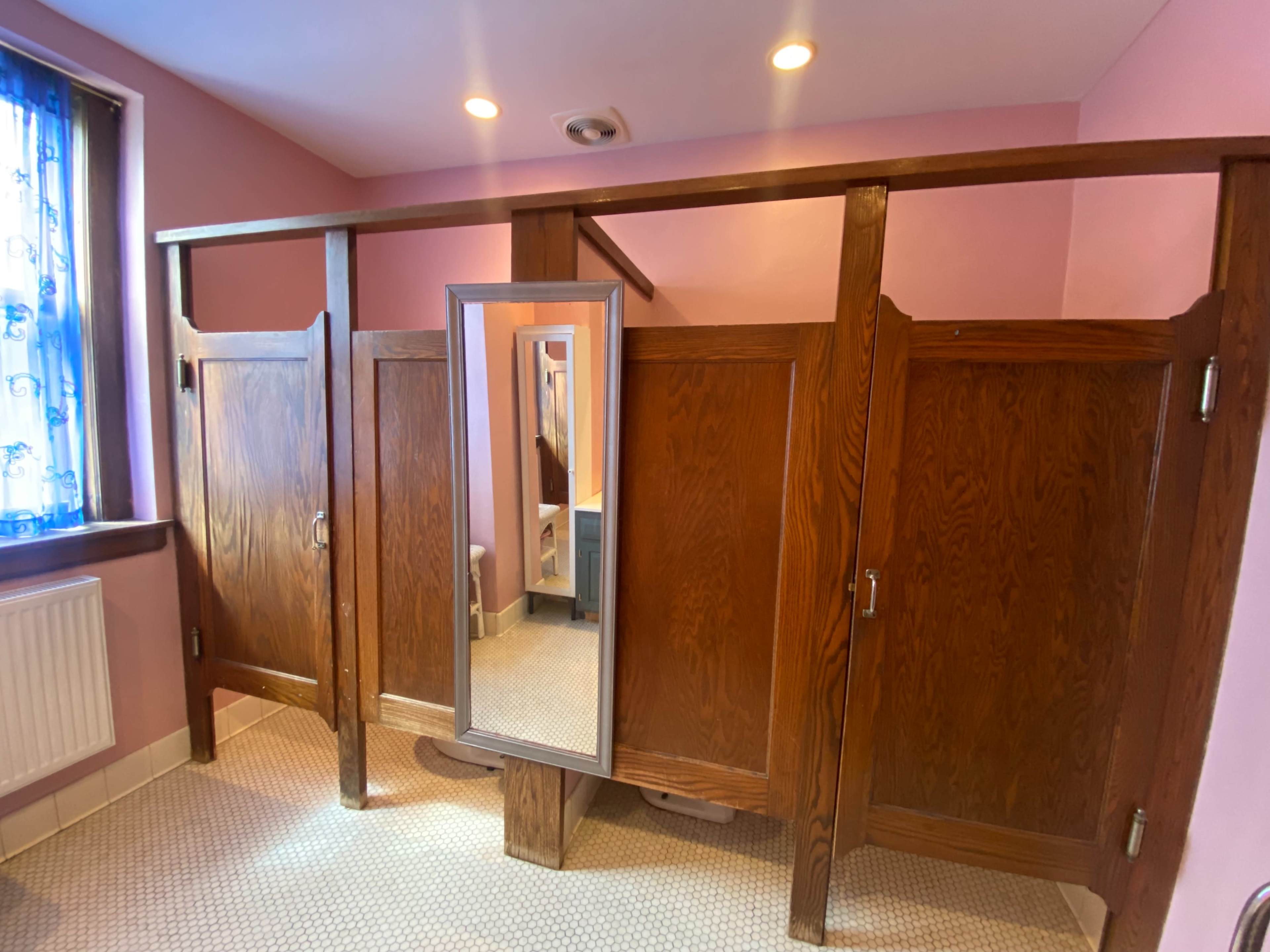 A restroom area with wooden stall partitions and a mirror, all set against a pink wall.