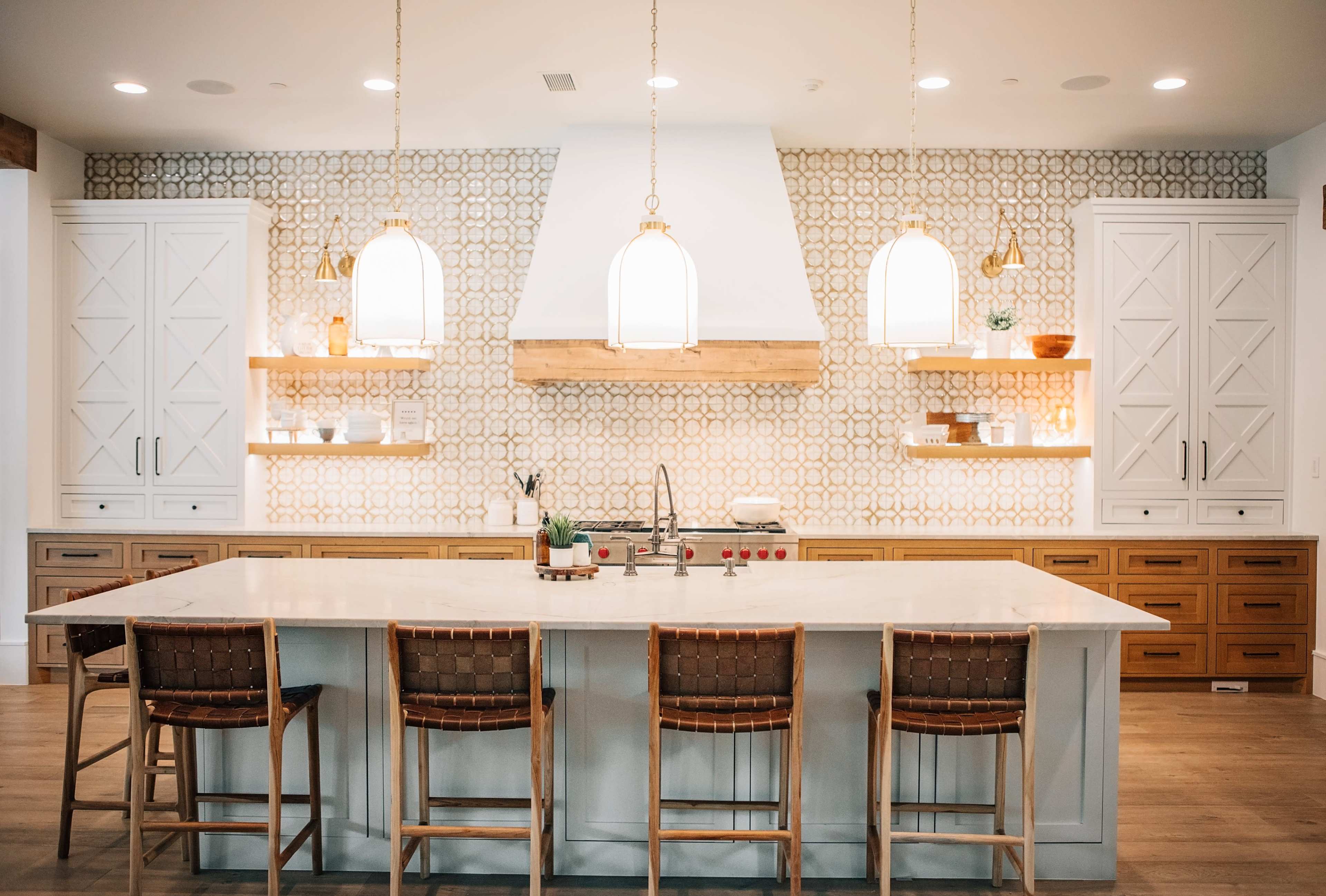 A modern kitchen features a large white island with wooden bar stools, decorative wall shelving, and patterned tile behind the stove.