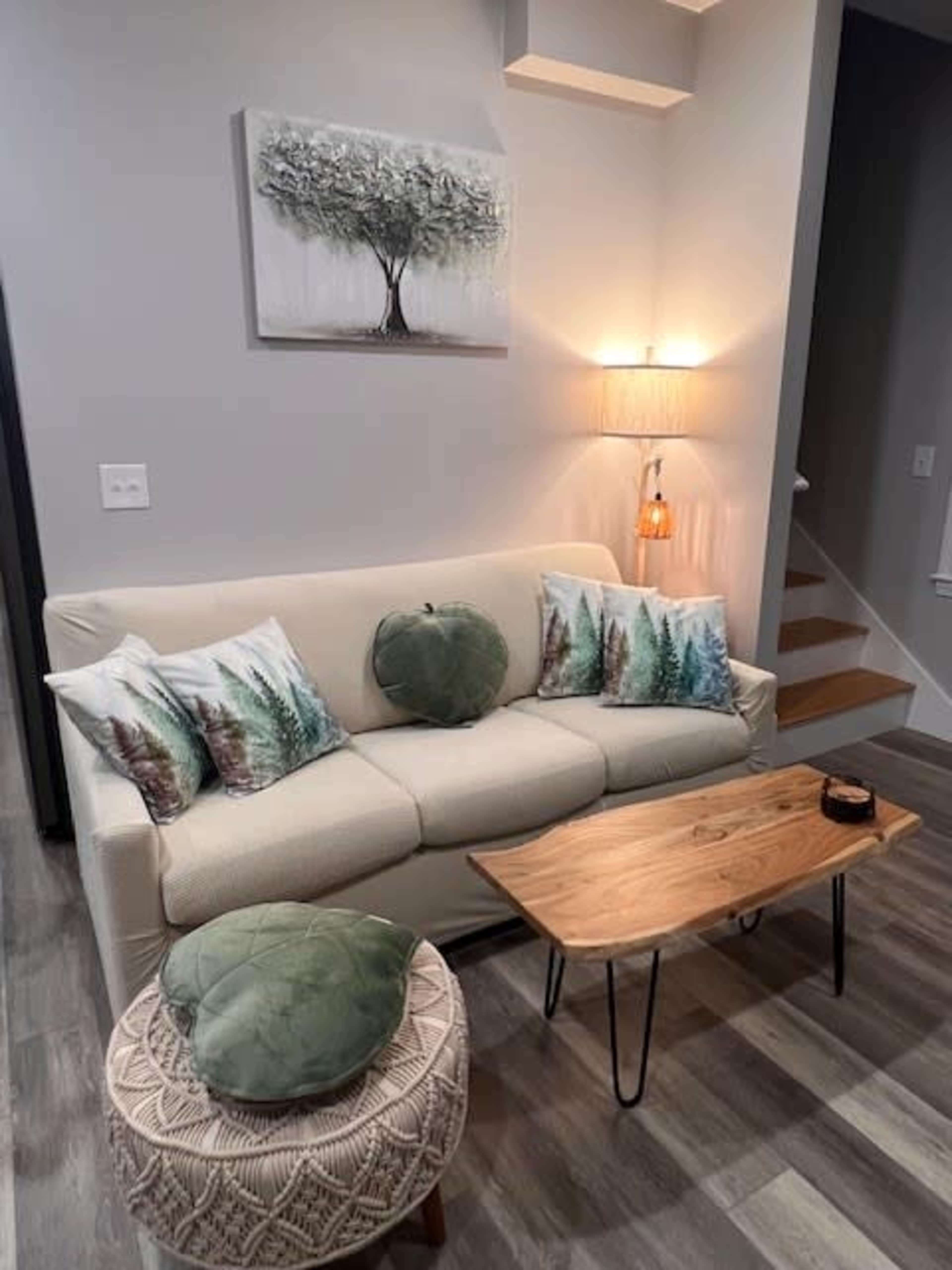 The image shows a living room with a light-colored sofa adorned with decorative cushions, a wooden coffee table, and a cozy armchair beside a staircase.
