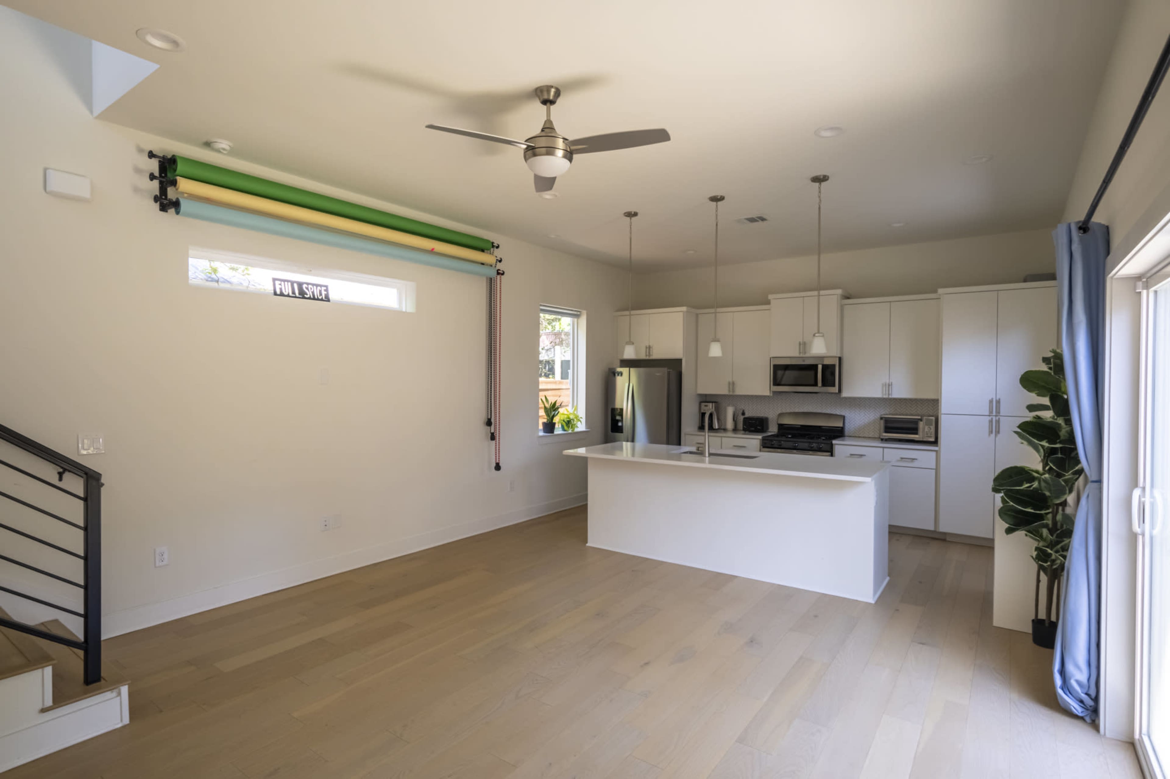 The image shows a modern kitchen and living area with light-colored walls, wooden flooring, and stainless steel appliances.