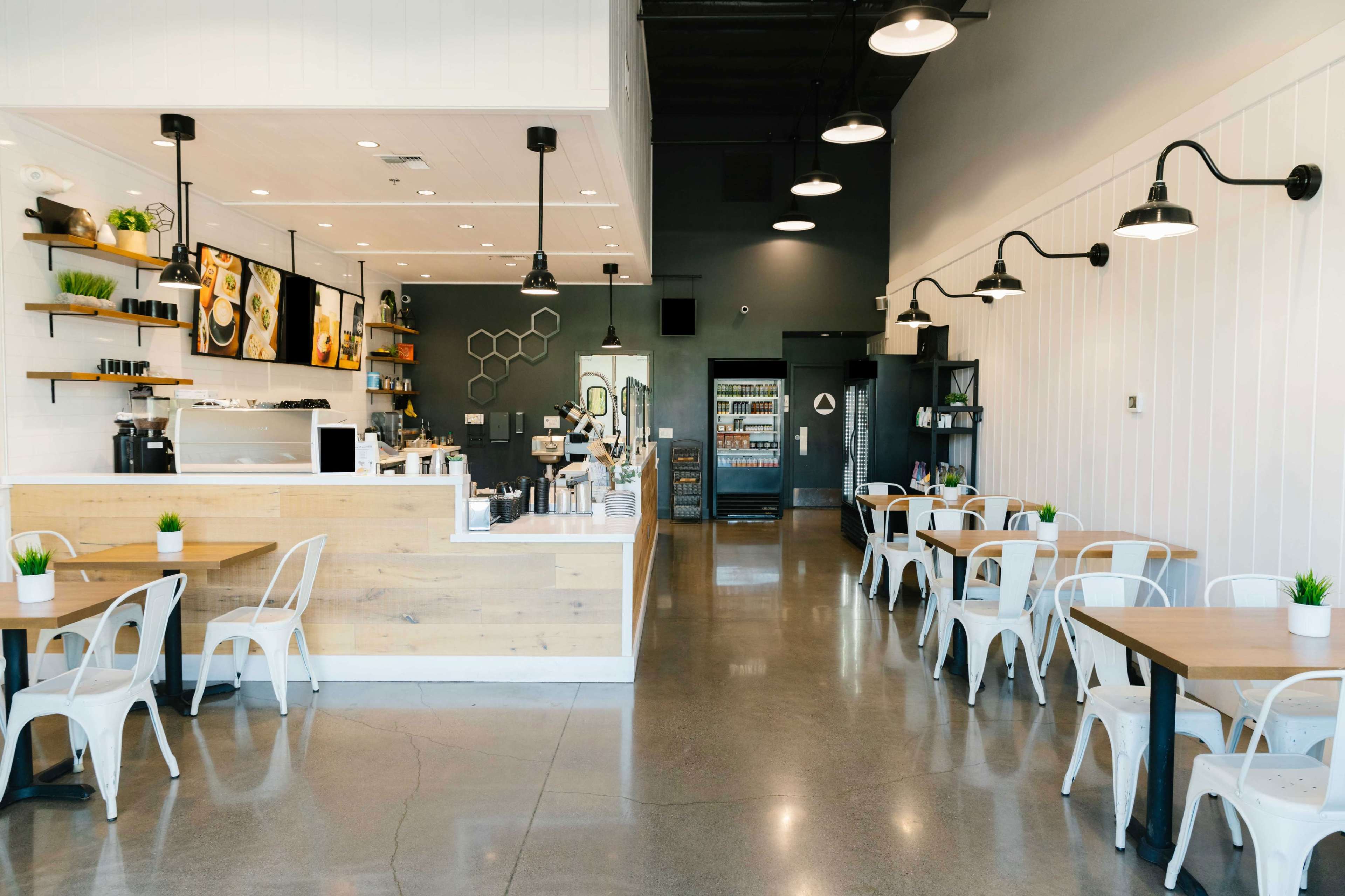 A modern café interior with wooden tables, white chairs, and a counter featuring coffee machines and displays.