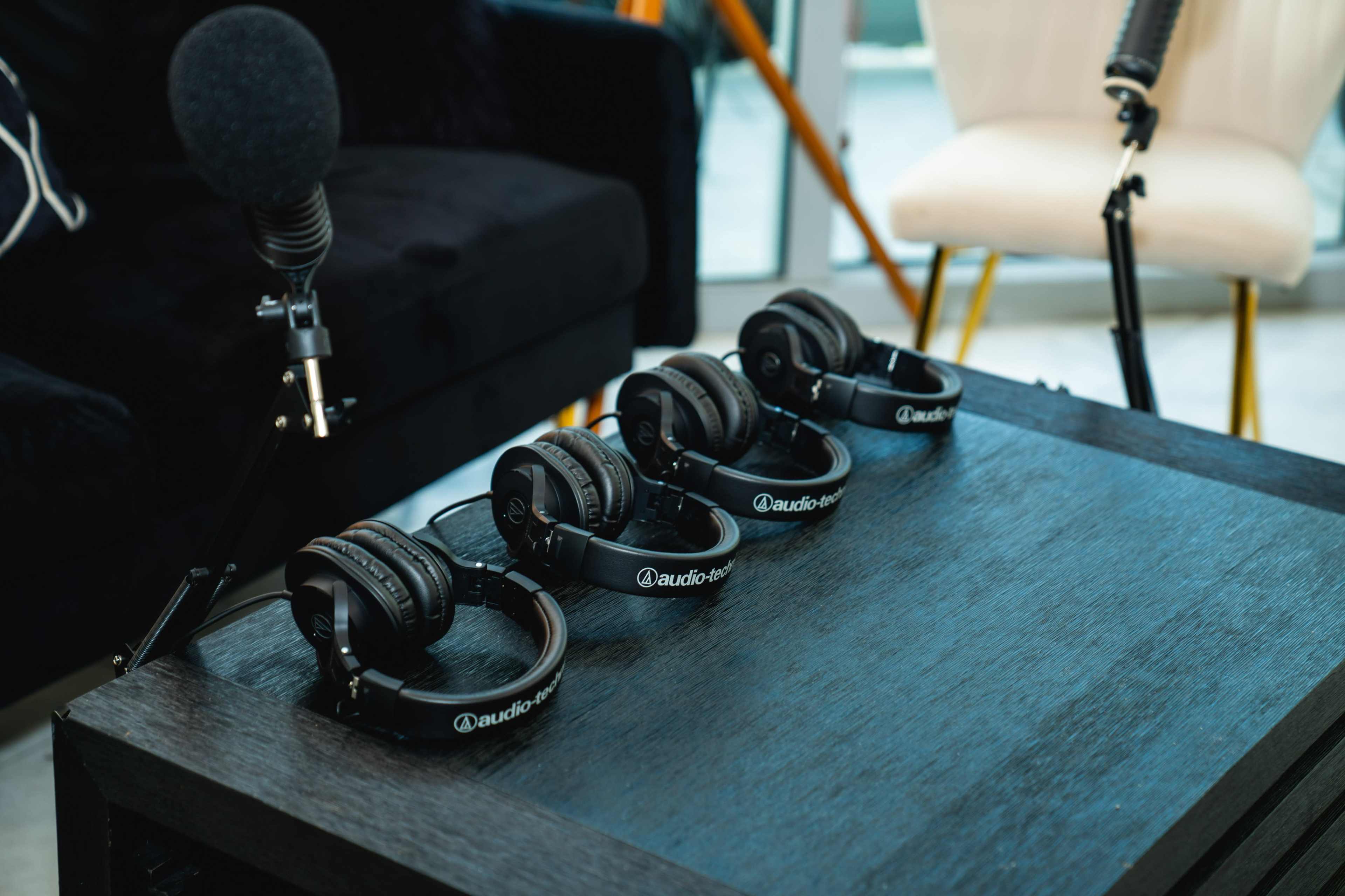 Four pairs of black headphones are laid out on a wooden table next to a microphone and a chair in a modern room.