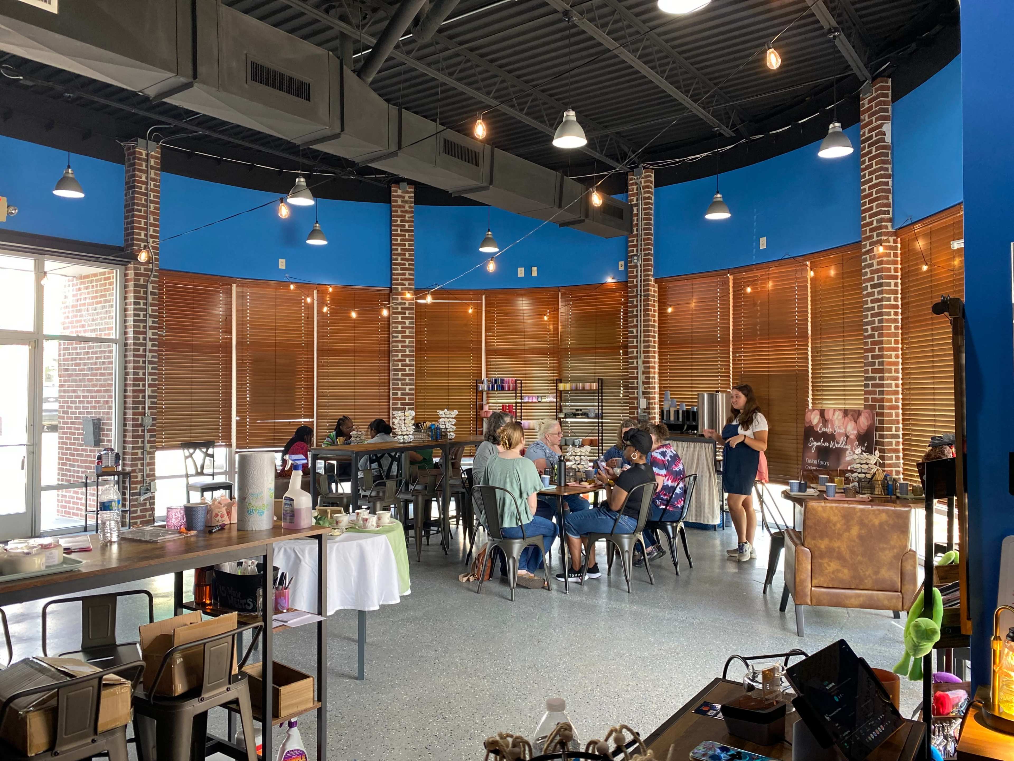 The image shows a brightly lit café with a circular seating arrangement, where several people are engaged in activities at tables, while others interact at a counter under string lights.