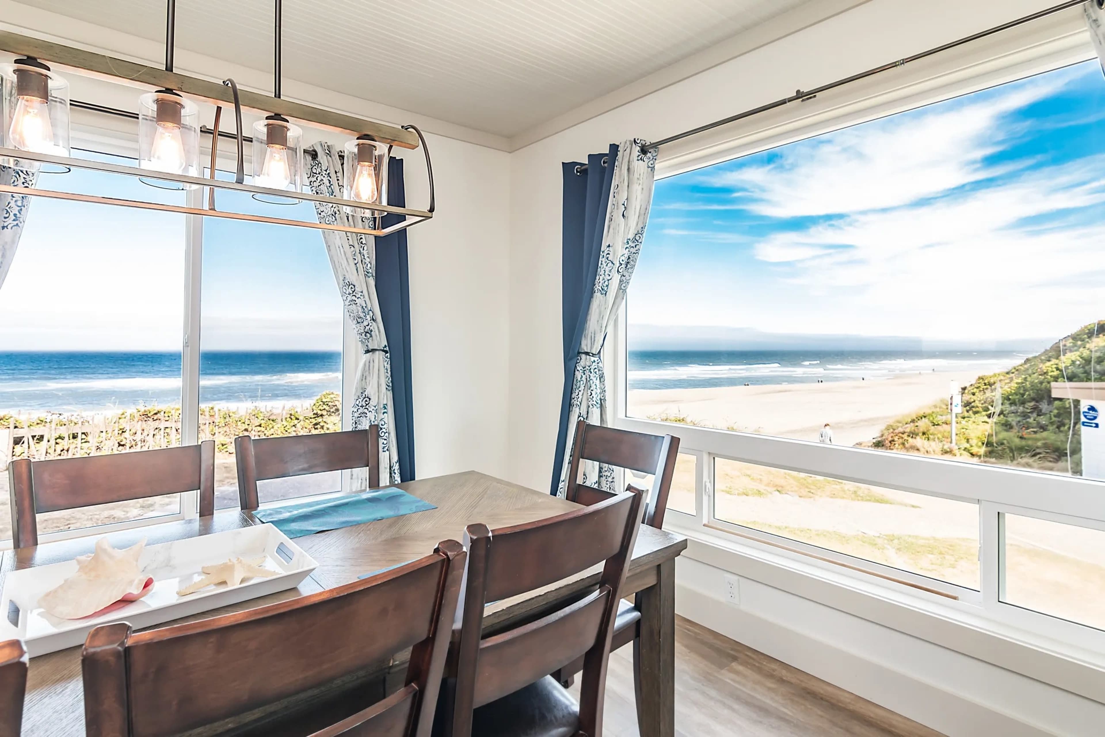 A dining area features a wooden table and chairs, with large windows offering a view of the ocean and beach.