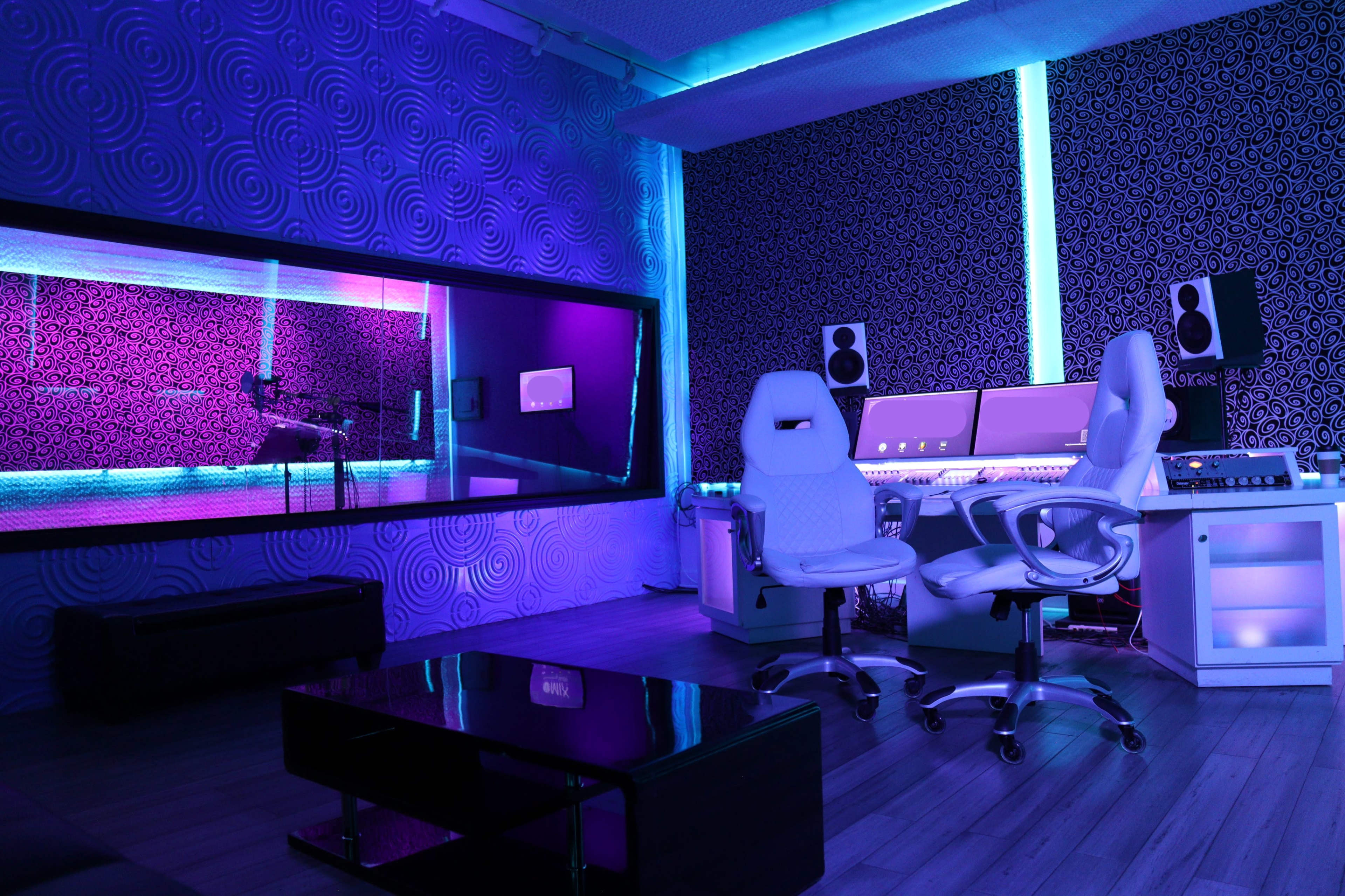 A modern recording studio with a microphone booth, two white chairs, and multi-colored LED lighting.