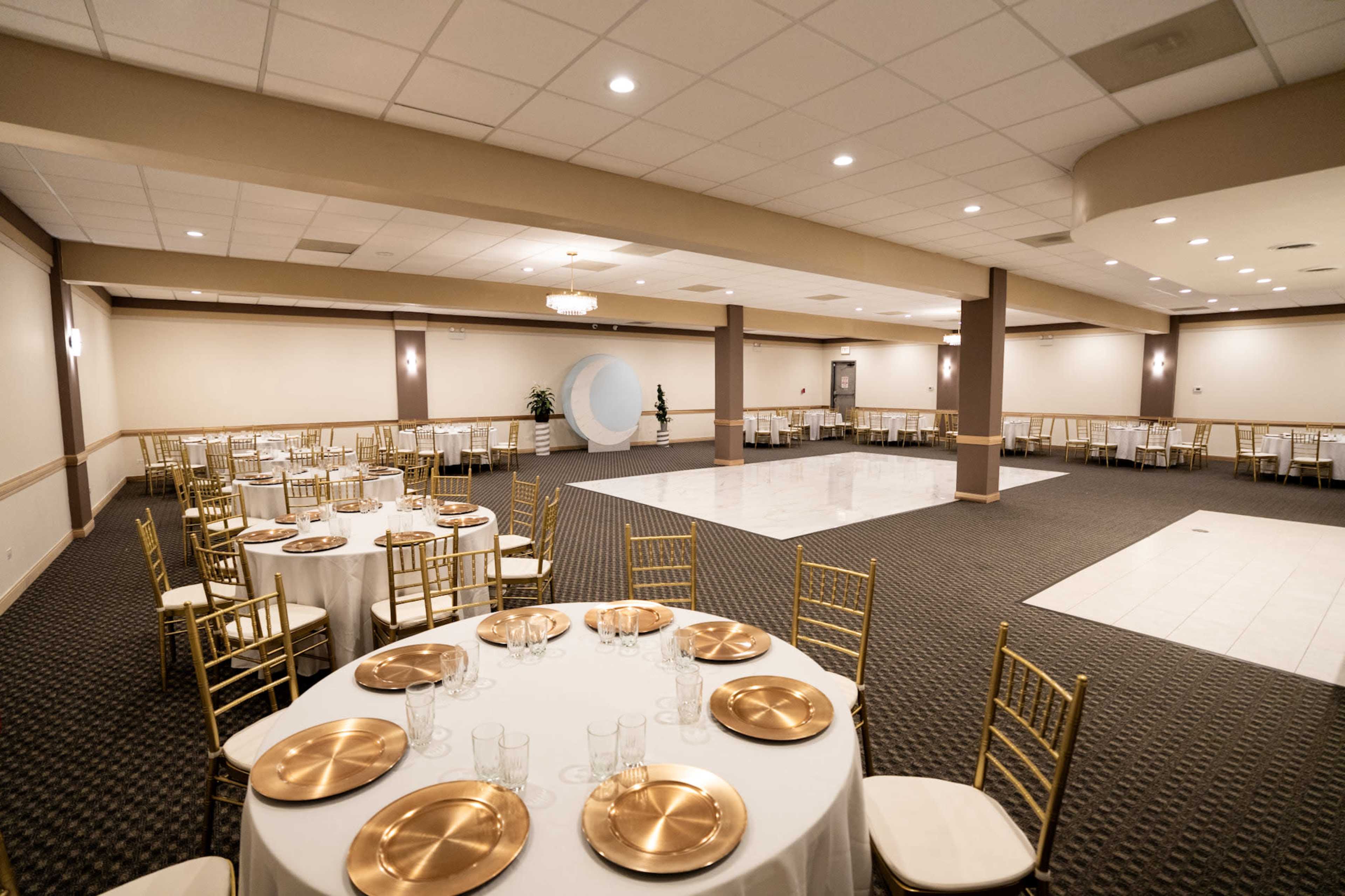 The image shows a banquet hall set up with round tables featuring gold plates and chairs, along with a large open space for dancing or activities.