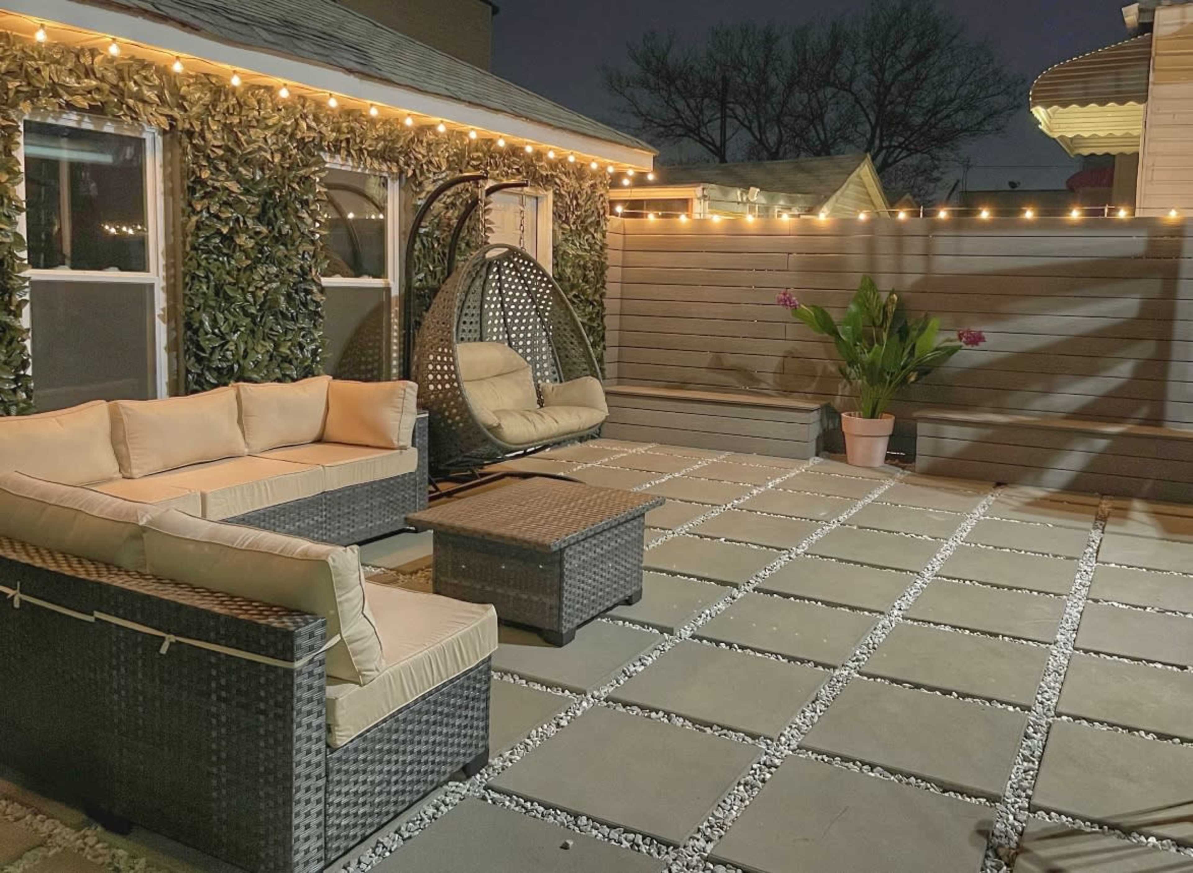 A cozy patio features a woven seating area with cushions, a hanging chair, and decorative lights illuminating the space at night.