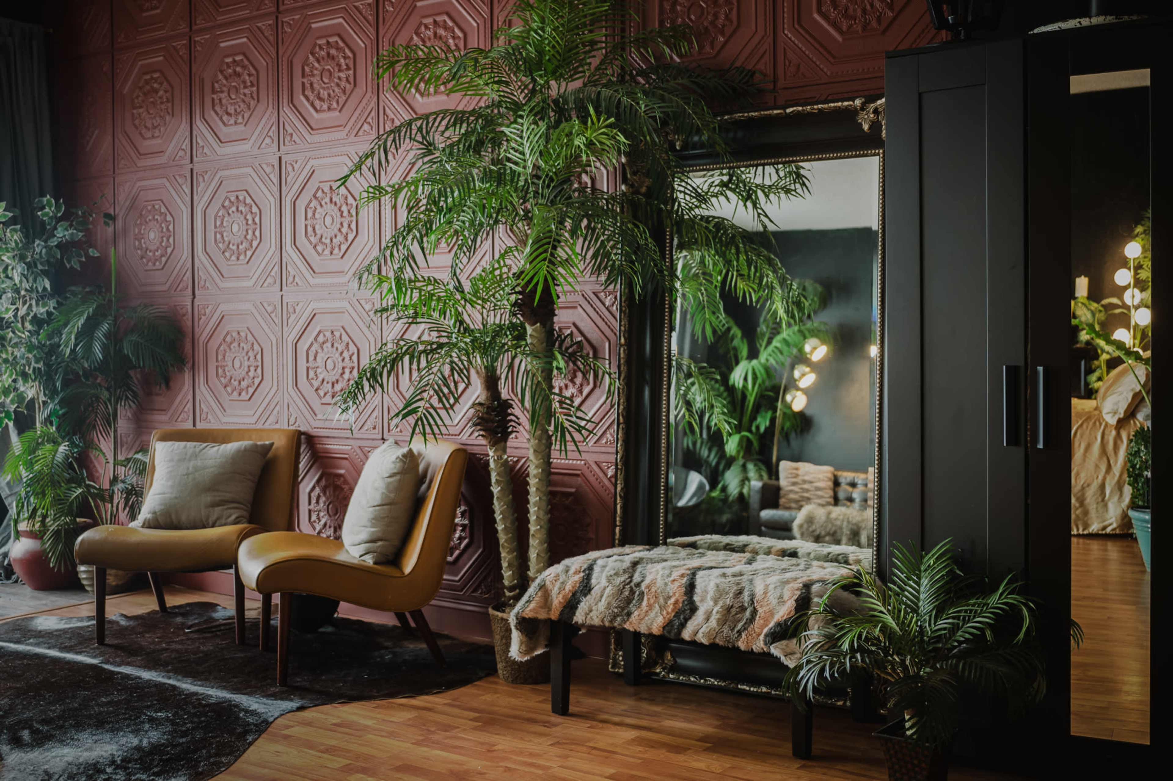 The image shows a stylish room featuring two armchairs, a mirrored wall, a plush bench, and decorative plants against a textured wallpaper.