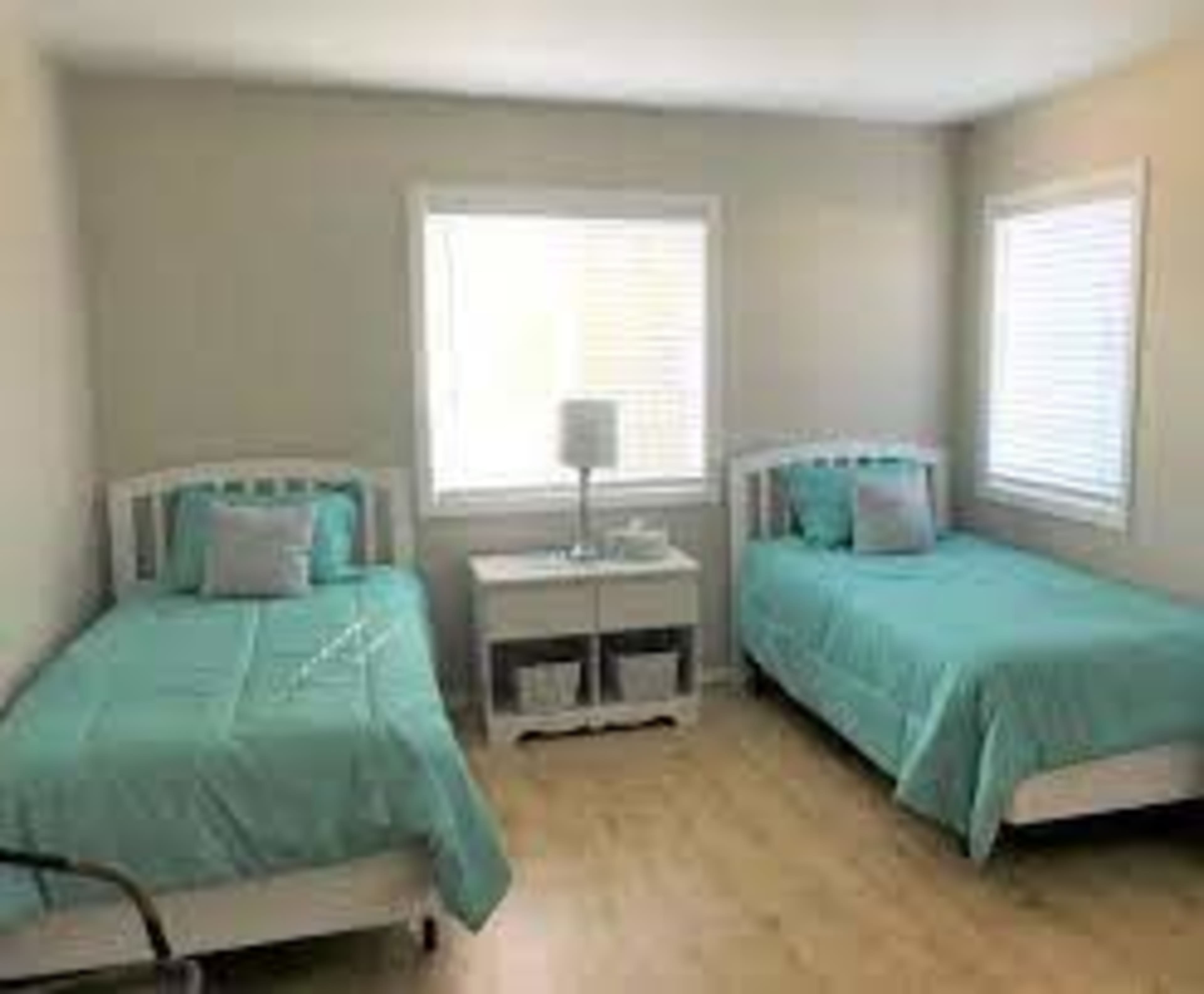The image shows a bedroom with two twin beds covered in turquoise bedding, a nightstand with a lamp between them, and windows allowing natural light.