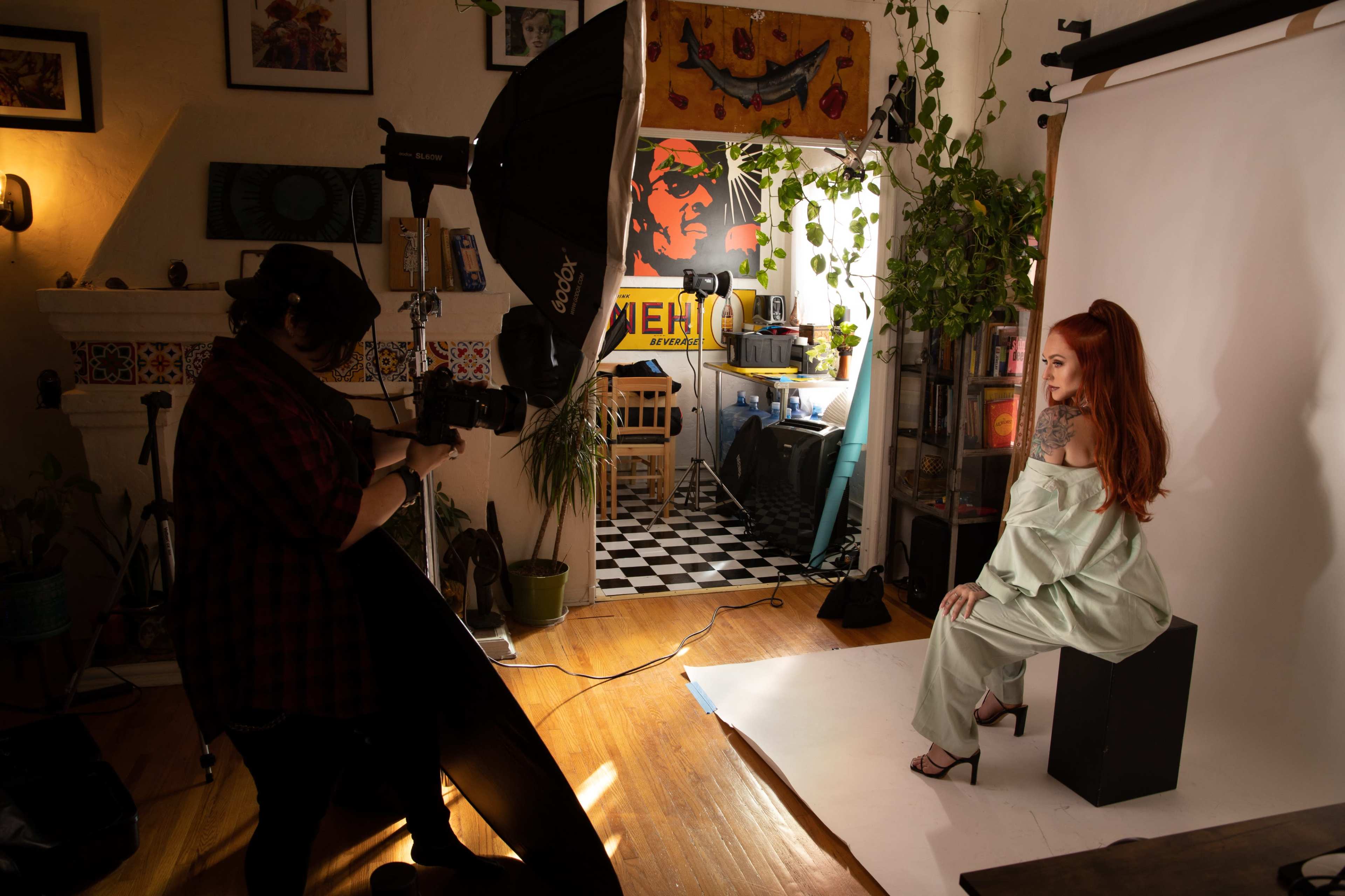 A photographer is taking a portrait of a woman seated on a stool in a well-decorated studio with a backdrop and natural light.