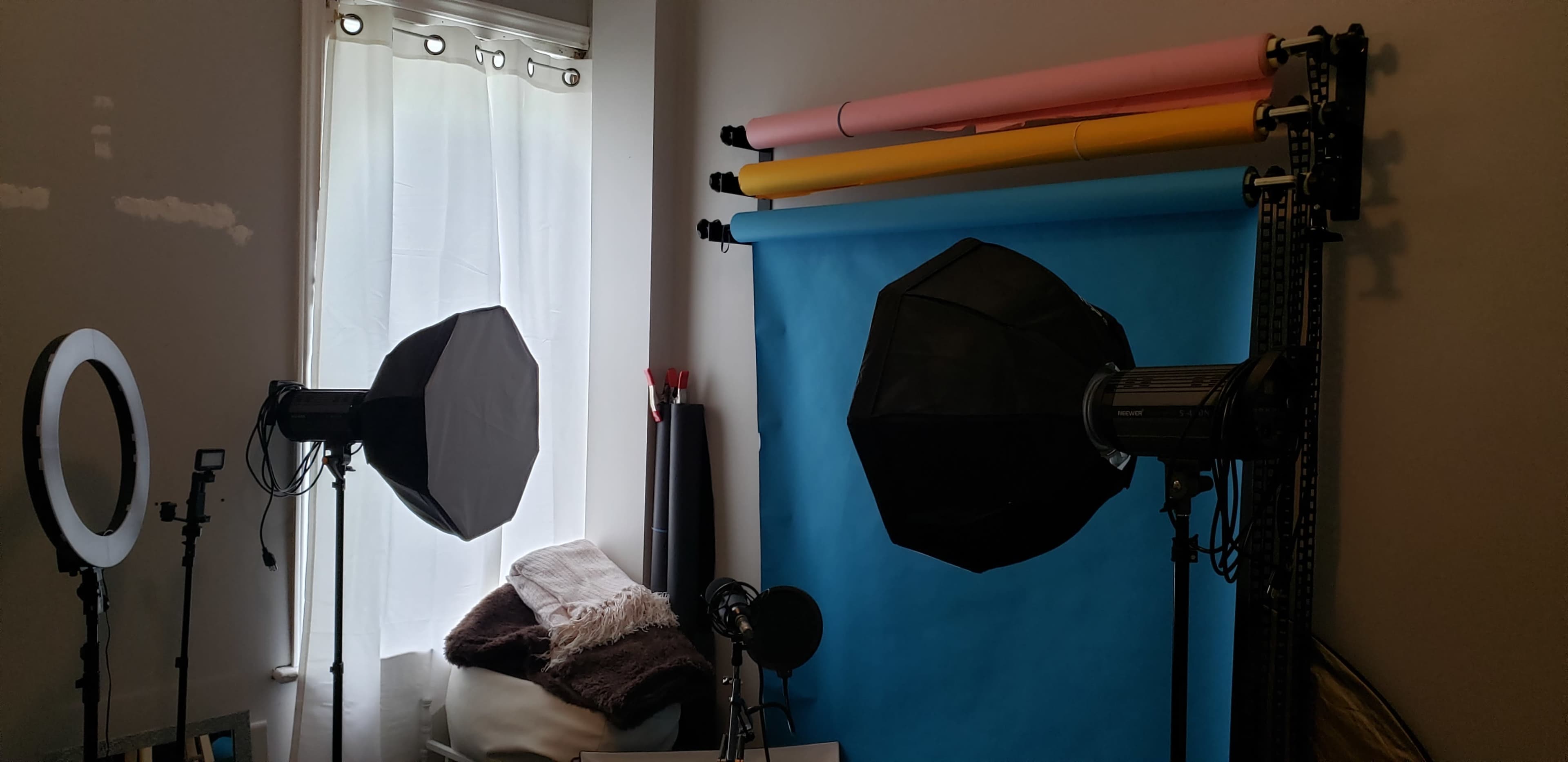 The image shows a photography setup with two softbox lights and a backdrop stand featuring rolls of colored paper against a wall, alongside a window with sheer curtains.
