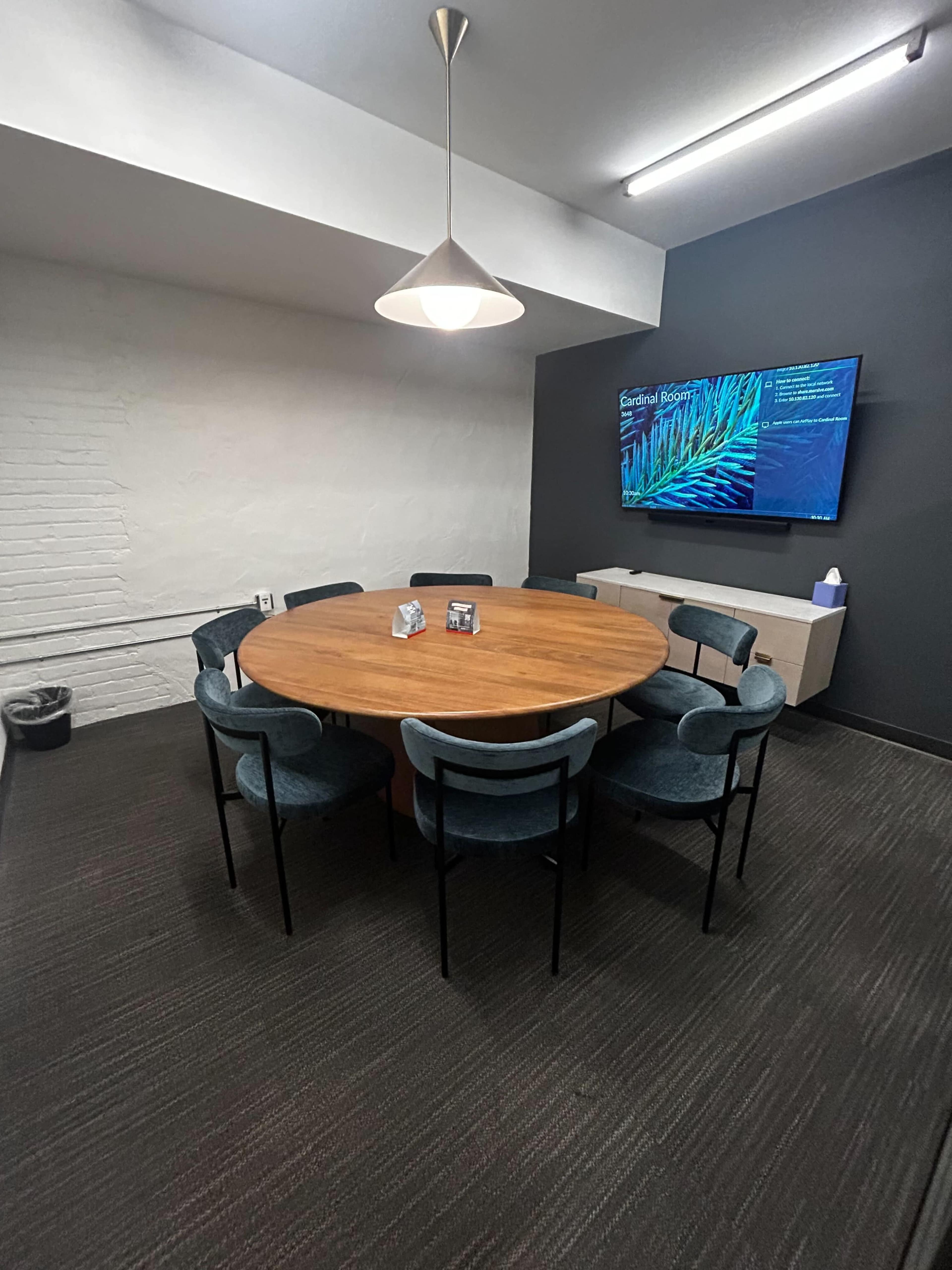 A circular wooden table surrounded by six upholstered chairs is set in a meeting room with a wall-mounted screen displaying information.
