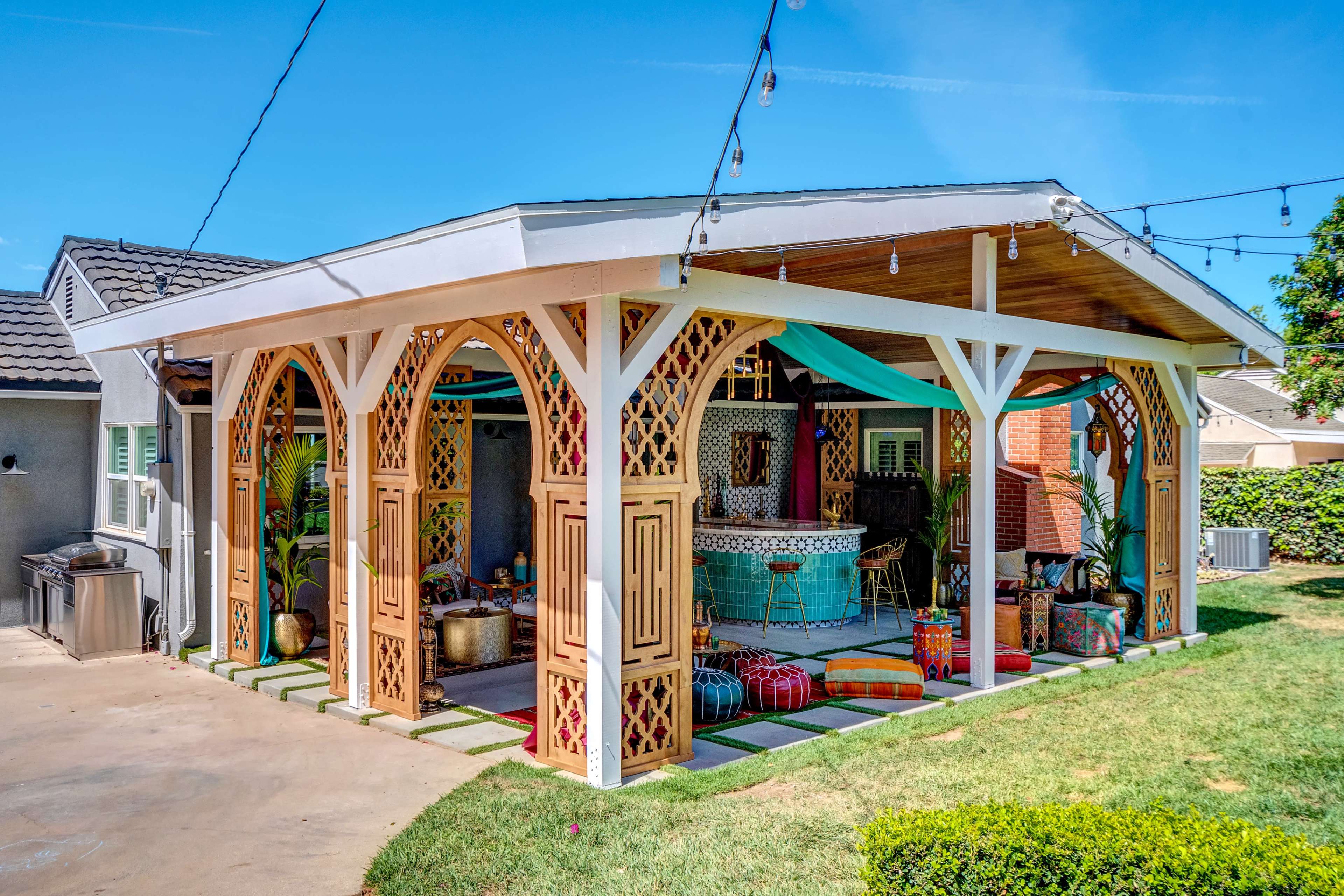 A spacious outdoor pavilion with decorative arches and colorful seating areas is set in a lush green yard.