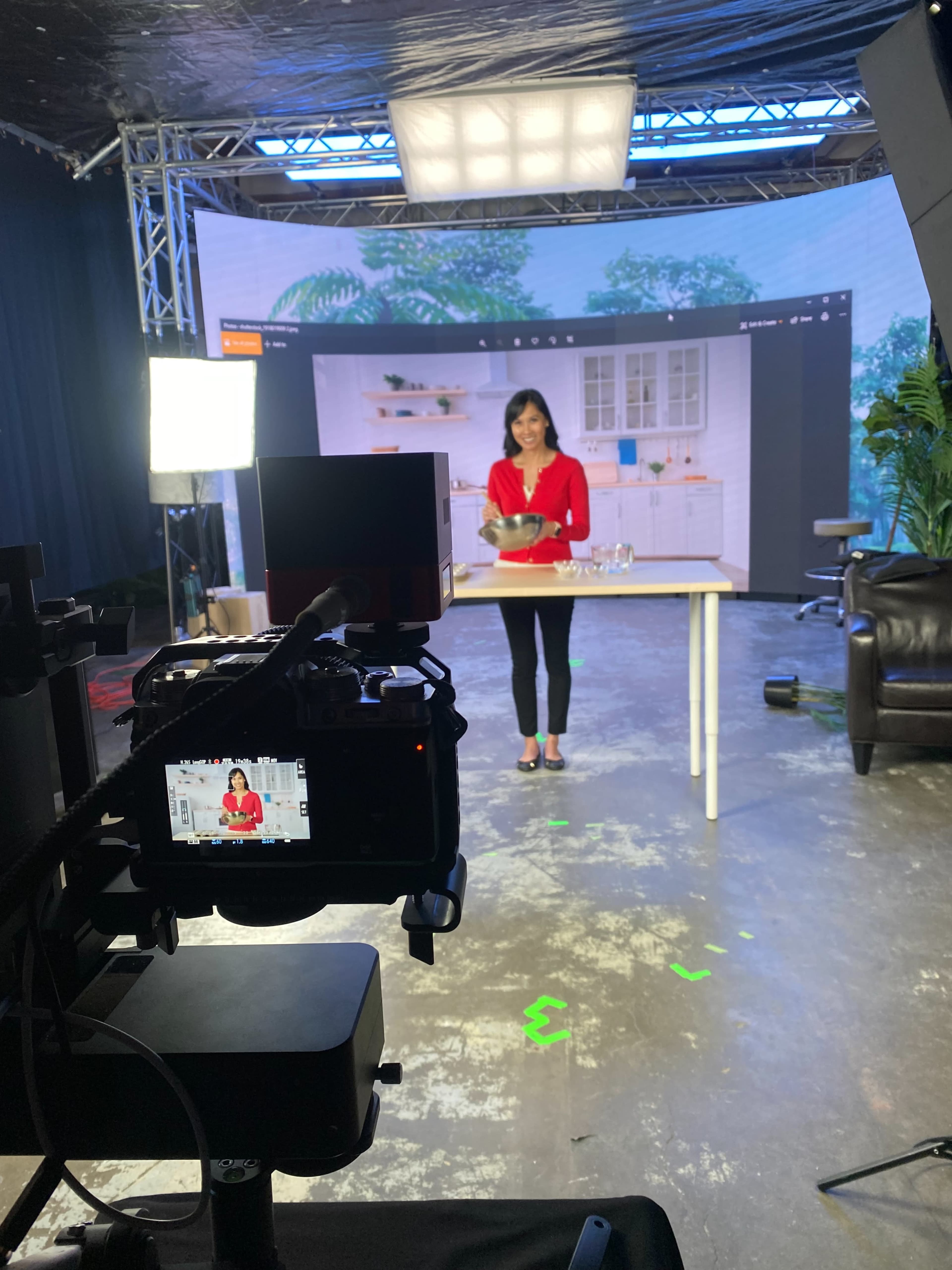 A woman in a red top stands behind a table in a studio setting, preparing to cook, while a camera focuses on her from the foreground.