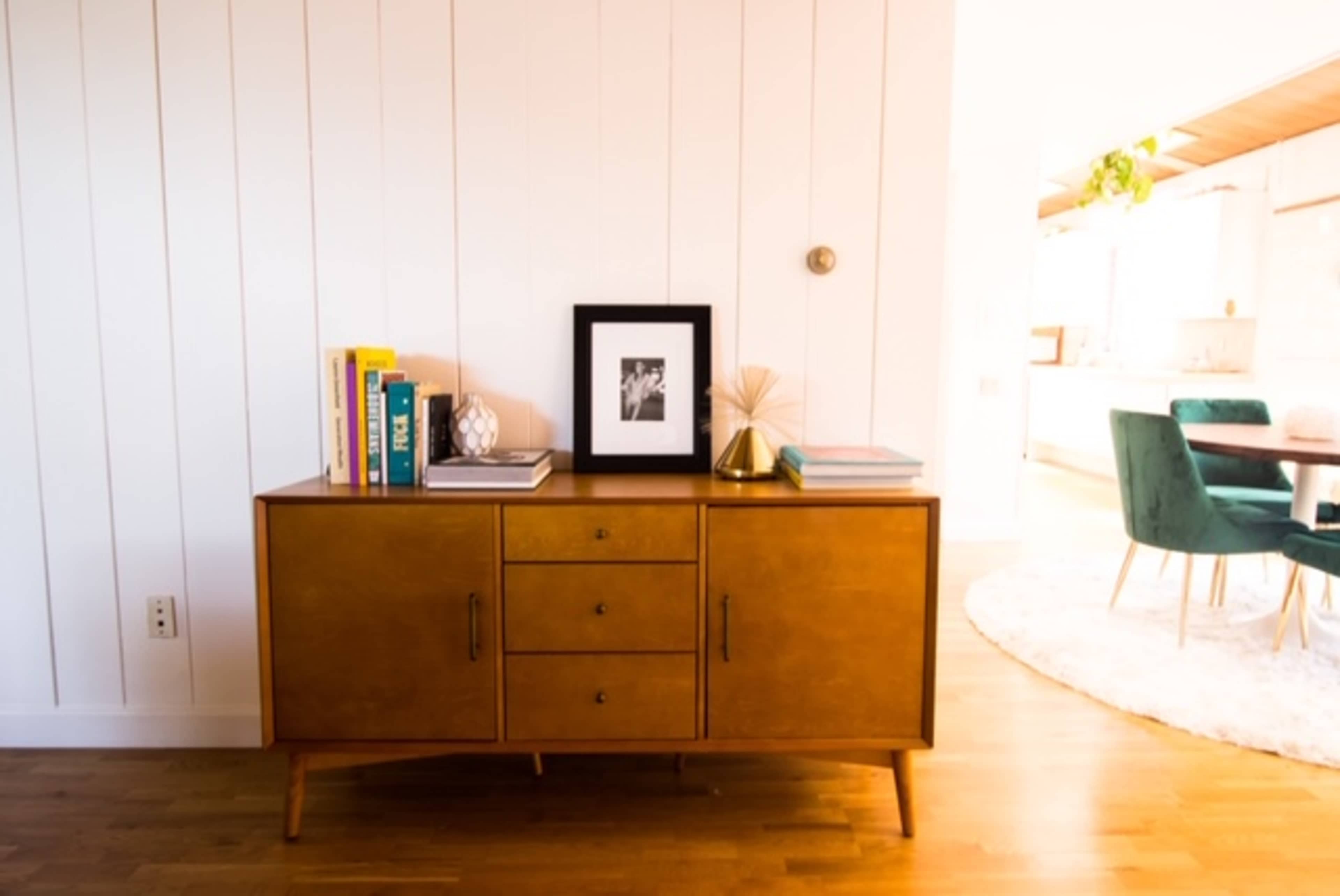 A mid-century modern wooden sideboard with three drawers and decorative items stands against a white-paneled wall, with a dining area visible in the background.