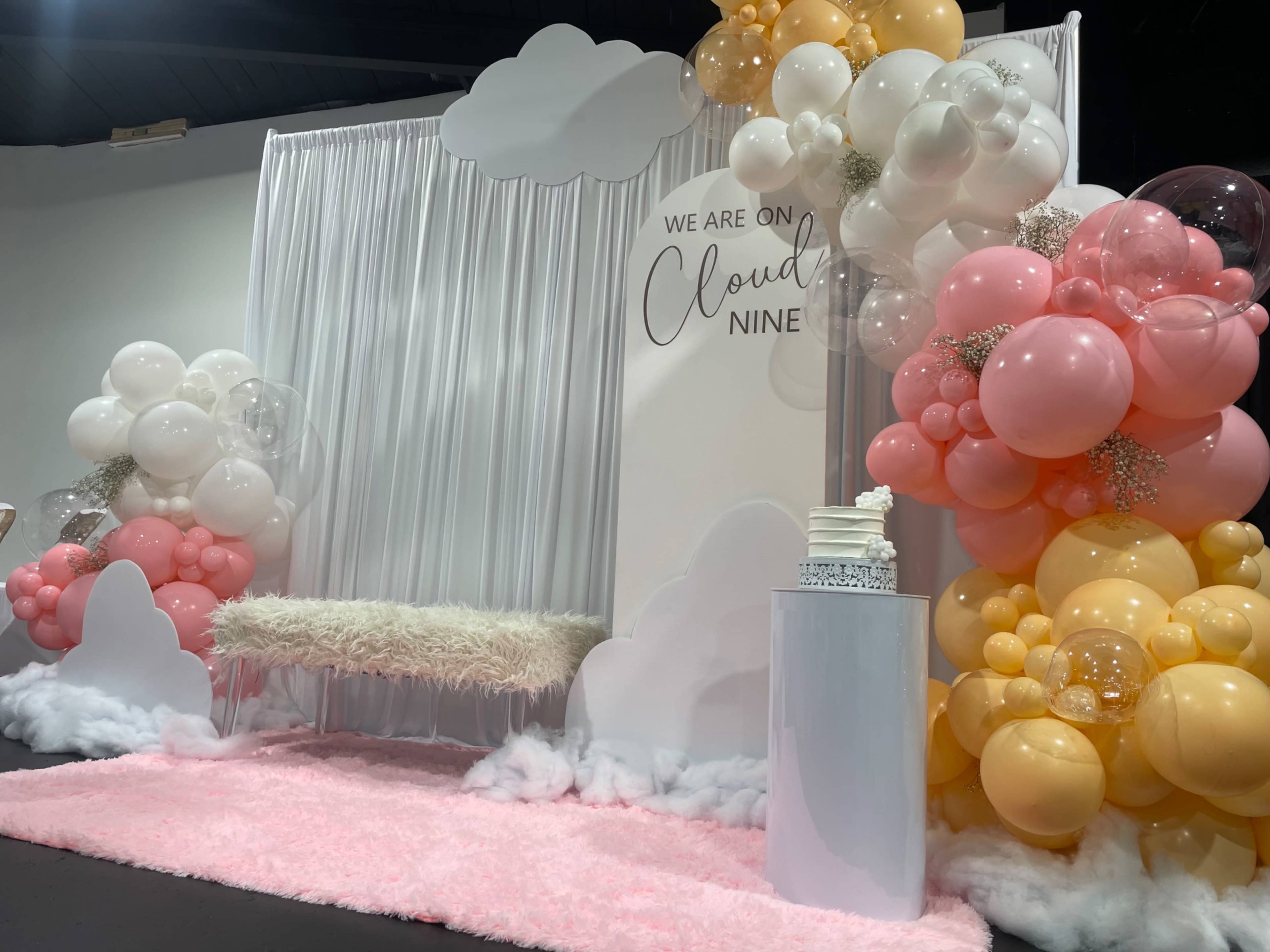 A photo of a decorative event backdrop featuring a fluffy white bench surrounded by colorful balloon arrangements and soft cloud decorations.