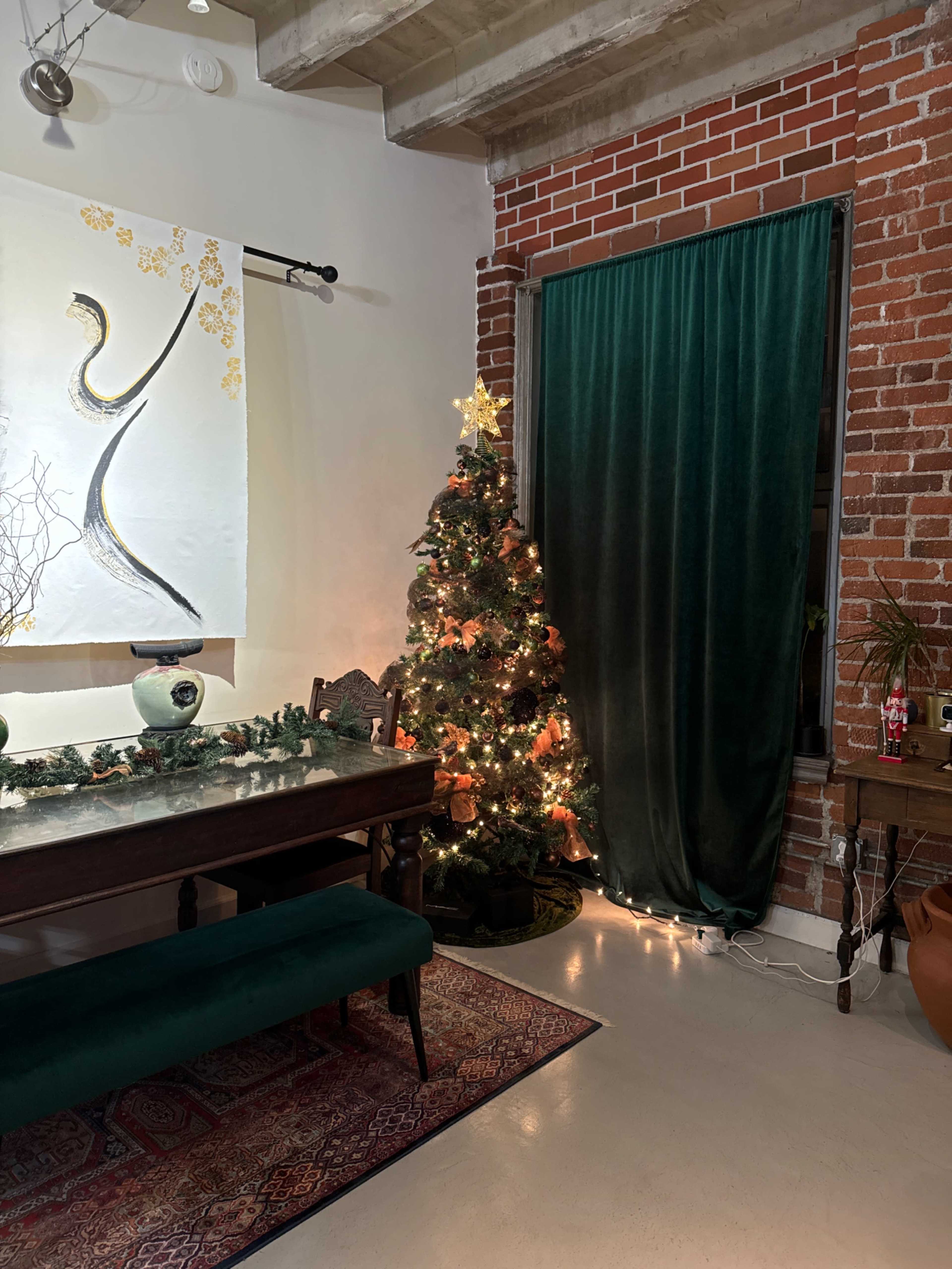 A decorated Christmas tree with lights and ornaments stands next to a green curtain in a room featuring a brick wall and a dining table.