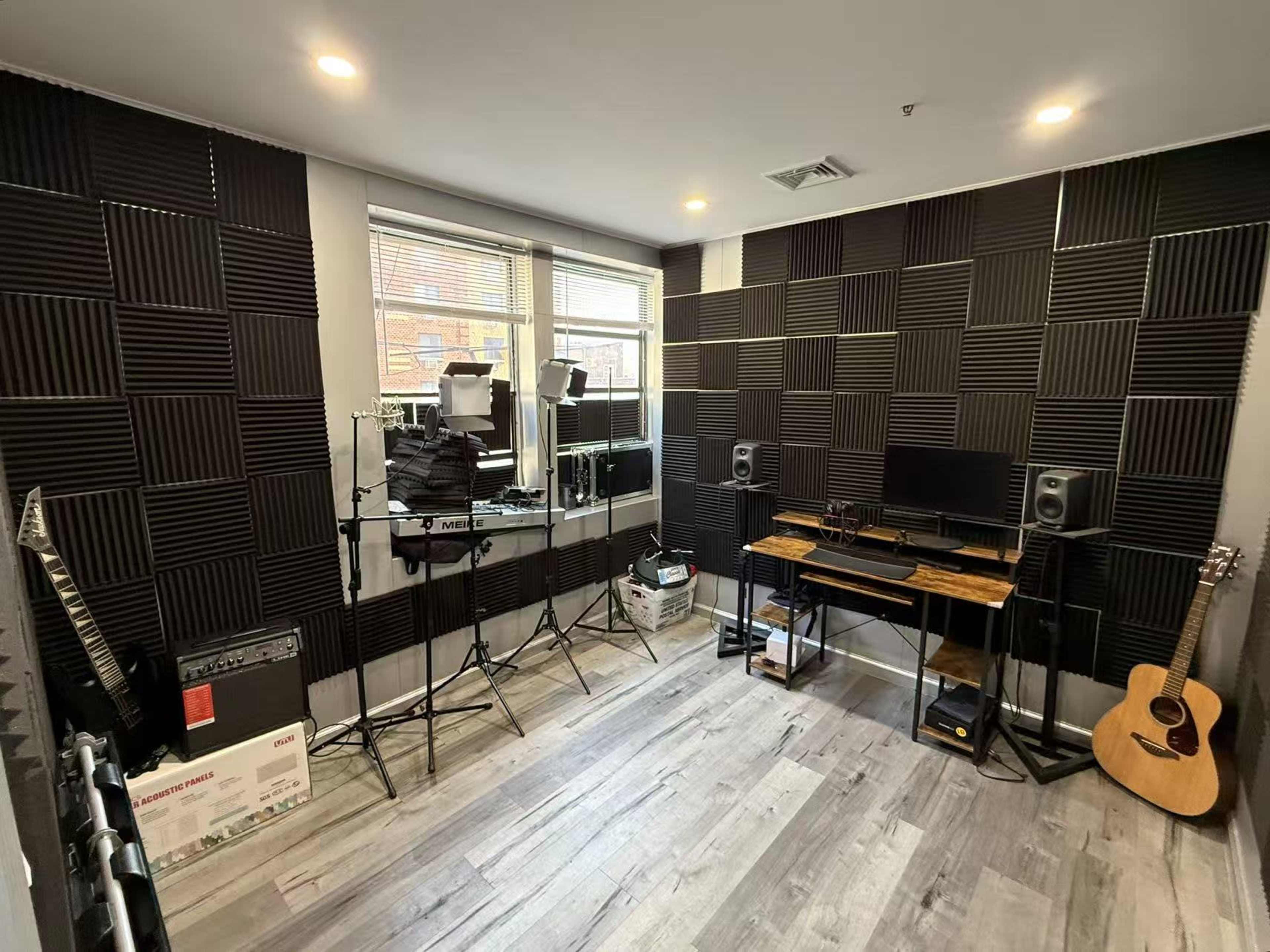 The image shows a recording studio with soundproof foam panels on the walls, studio lights on stands, a computer setup, and an acoustic guitar in the corner.
