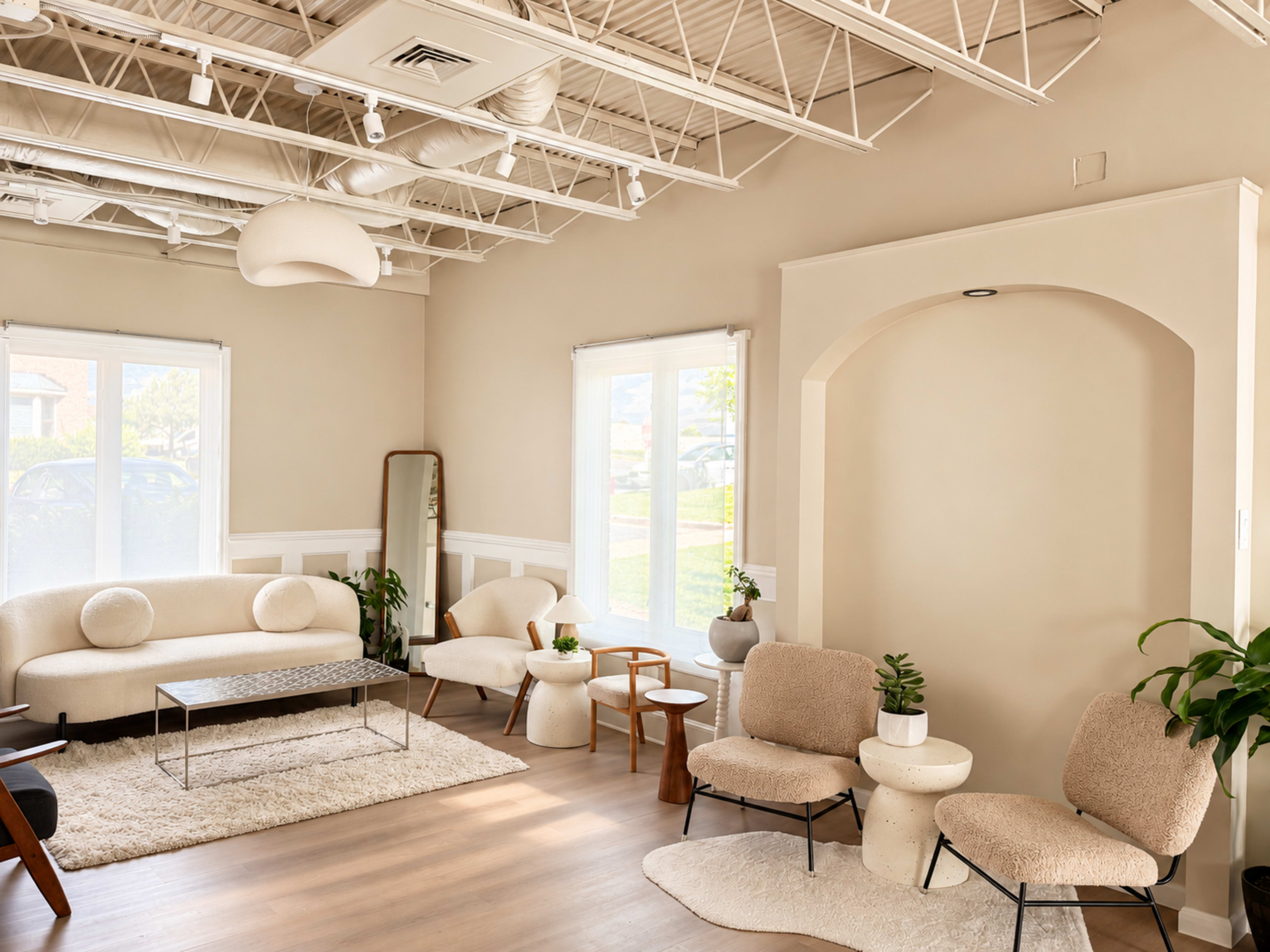 The image shows a modern living room with neutral-colored walls, a large sofa, two upholstered chairs, and a coffee table surrounded by plants and natural light from windows.