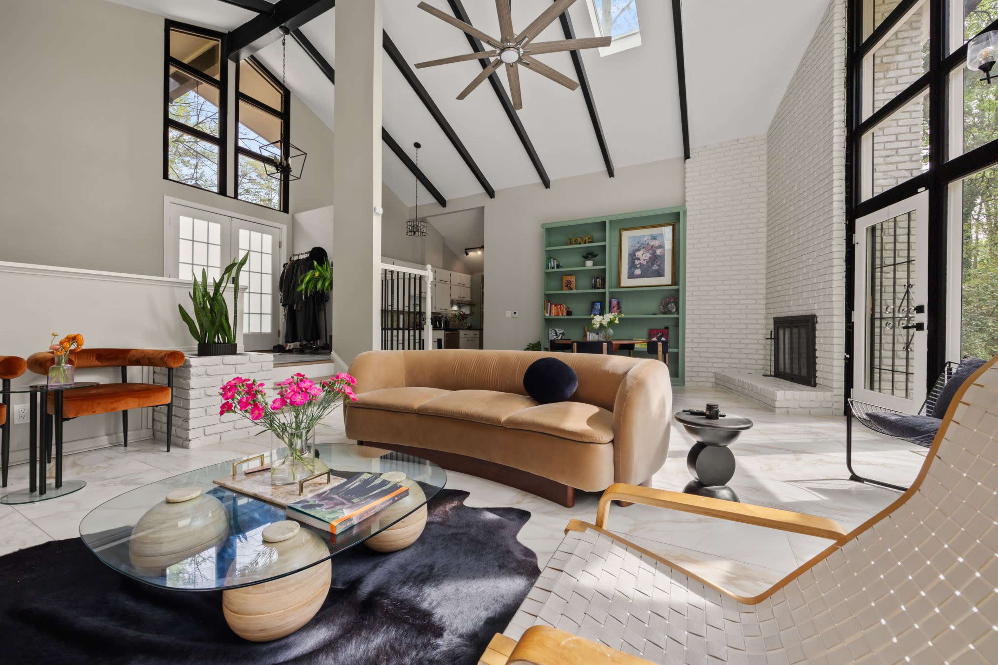 The image shows a modern living room with high ceilings, large windows, and a mix of contemporary furniture including a curved sofa, a coffee table, and a cozy armchair.