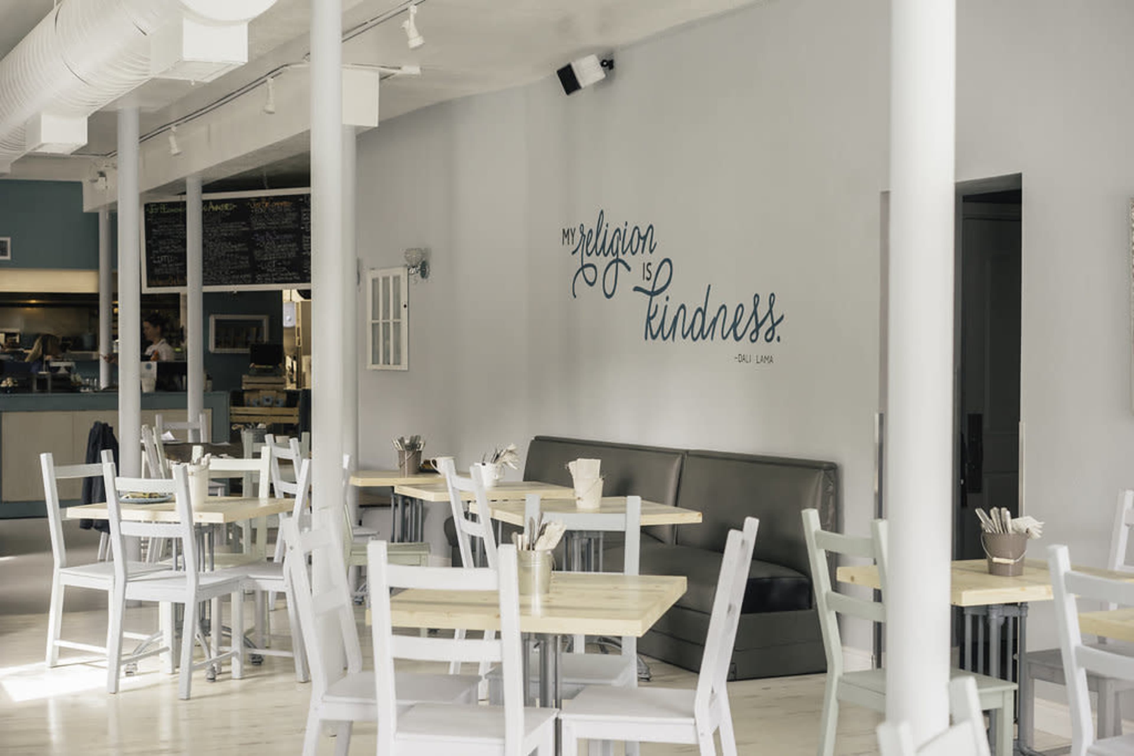 The interior of a café features simple wooden tables and chairs, with a wall displaying the inscription "my religion is kindness."