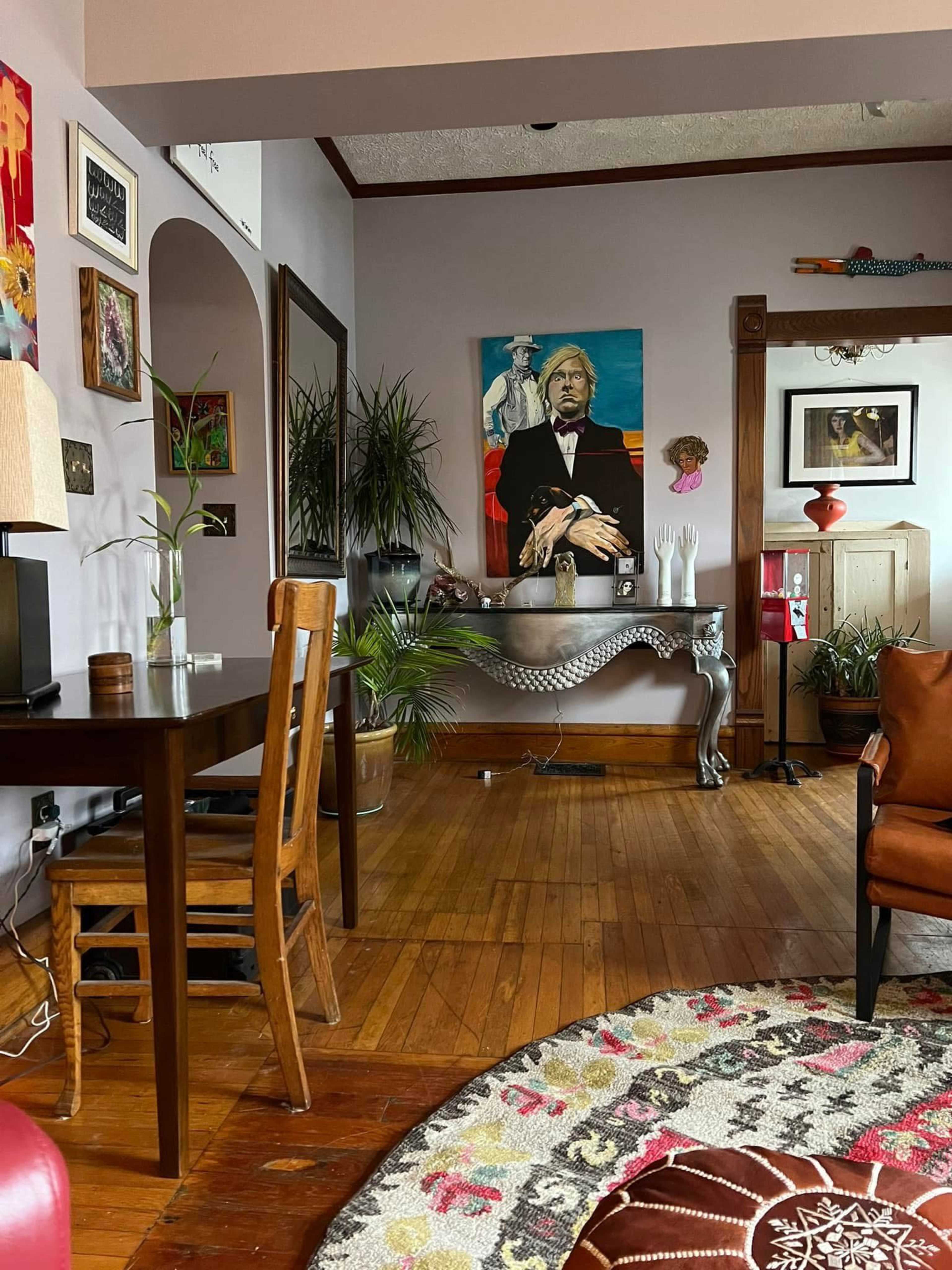 A wooden table and chair are positioned in a cozy living room, featuring a large painting on the wall and various plants throughout the space.
