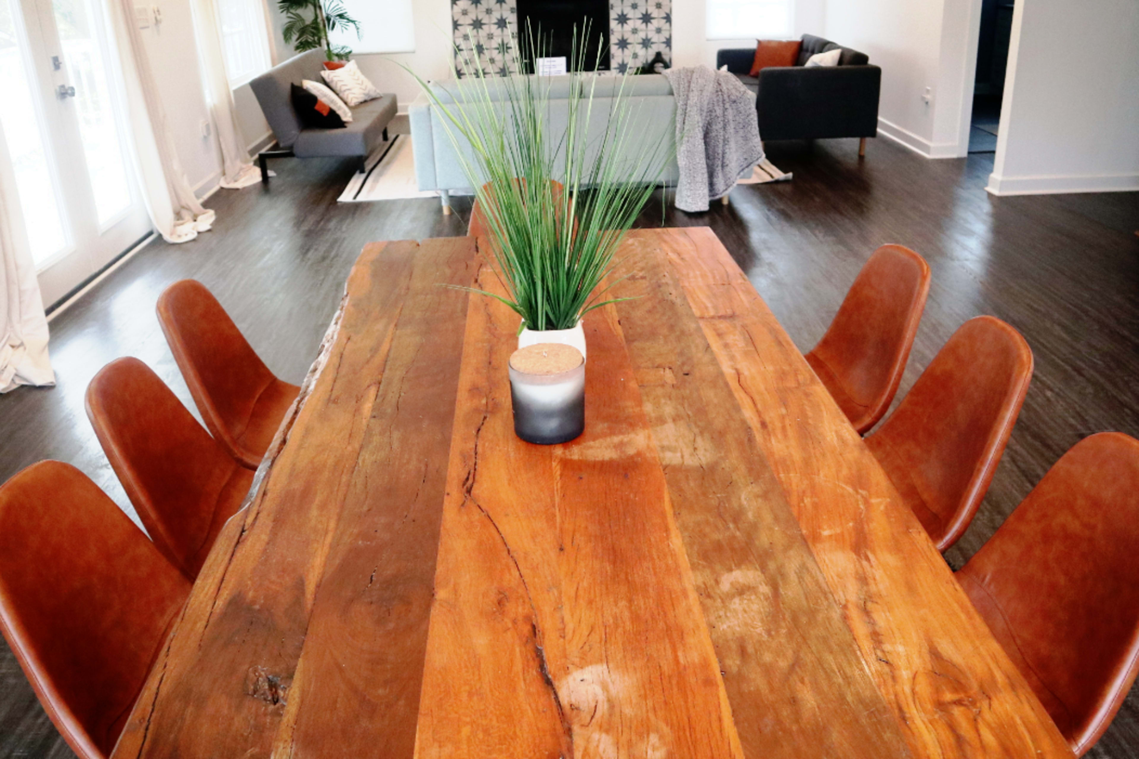 A wooden dining table with brown leather chairs surrounds it, featuring a small potted plant in the center, and a cozy living area visible in the background.