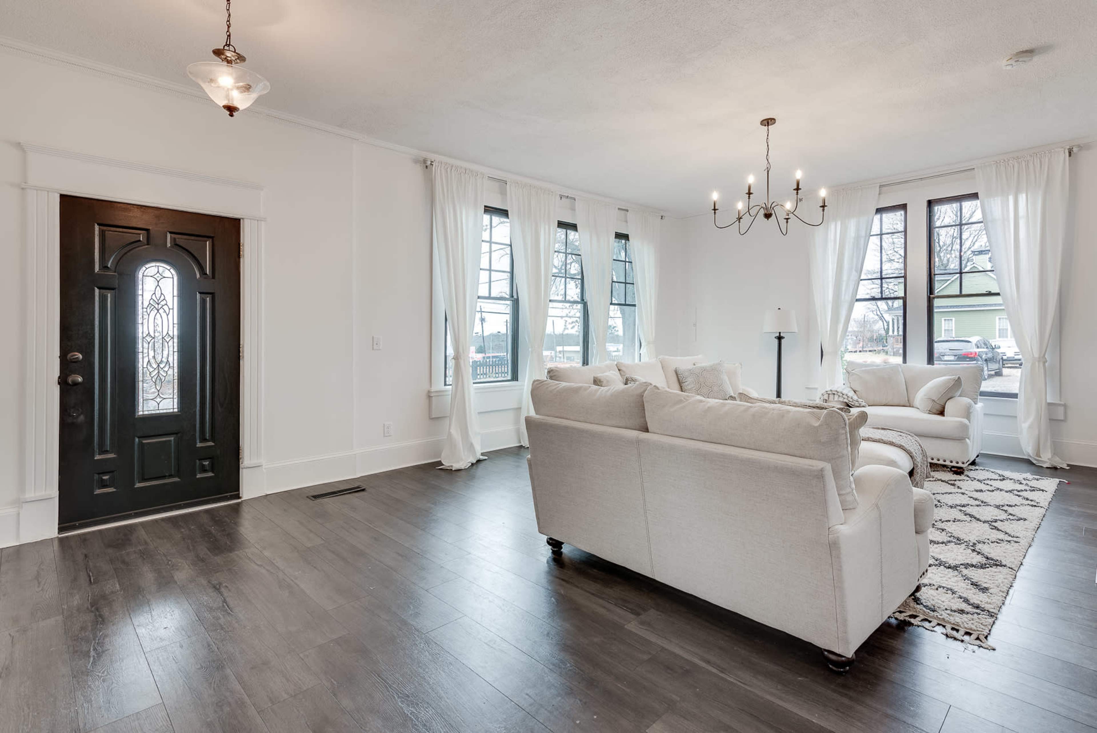 A well-lit living room featuring a white sofa set, large windows, and a black front door.