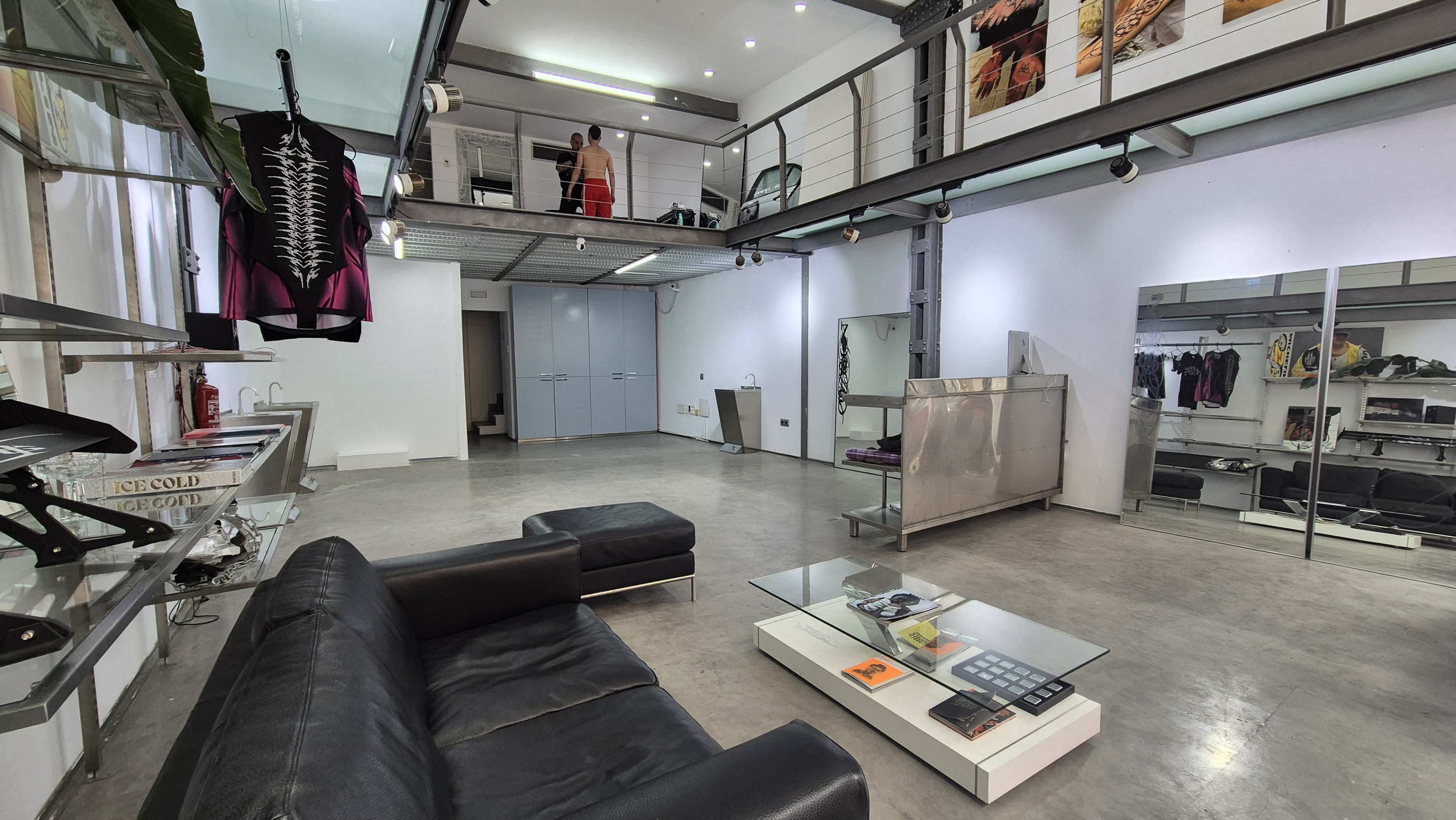 The image shows a modern, spacious interior with a seating area, a glass coffee table, and various clothing displays arranged on the walls.