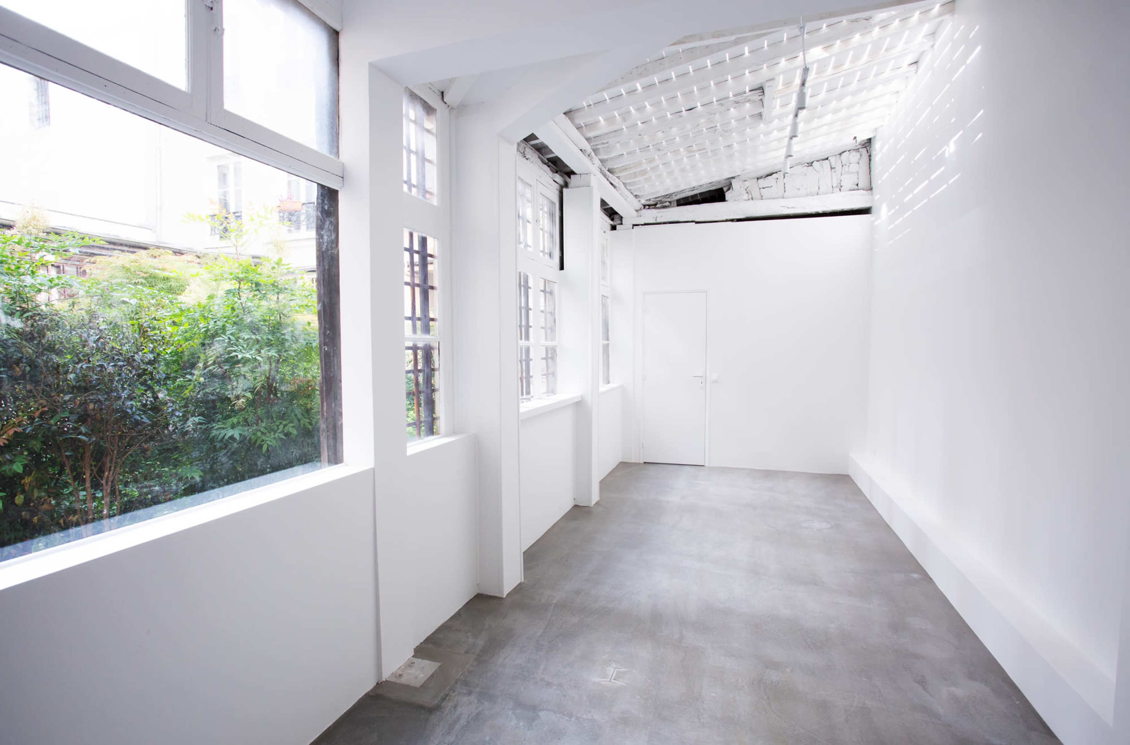 The image shows a spacious, empty room with white walls, large windows allowing natural light, and a concrete floor.