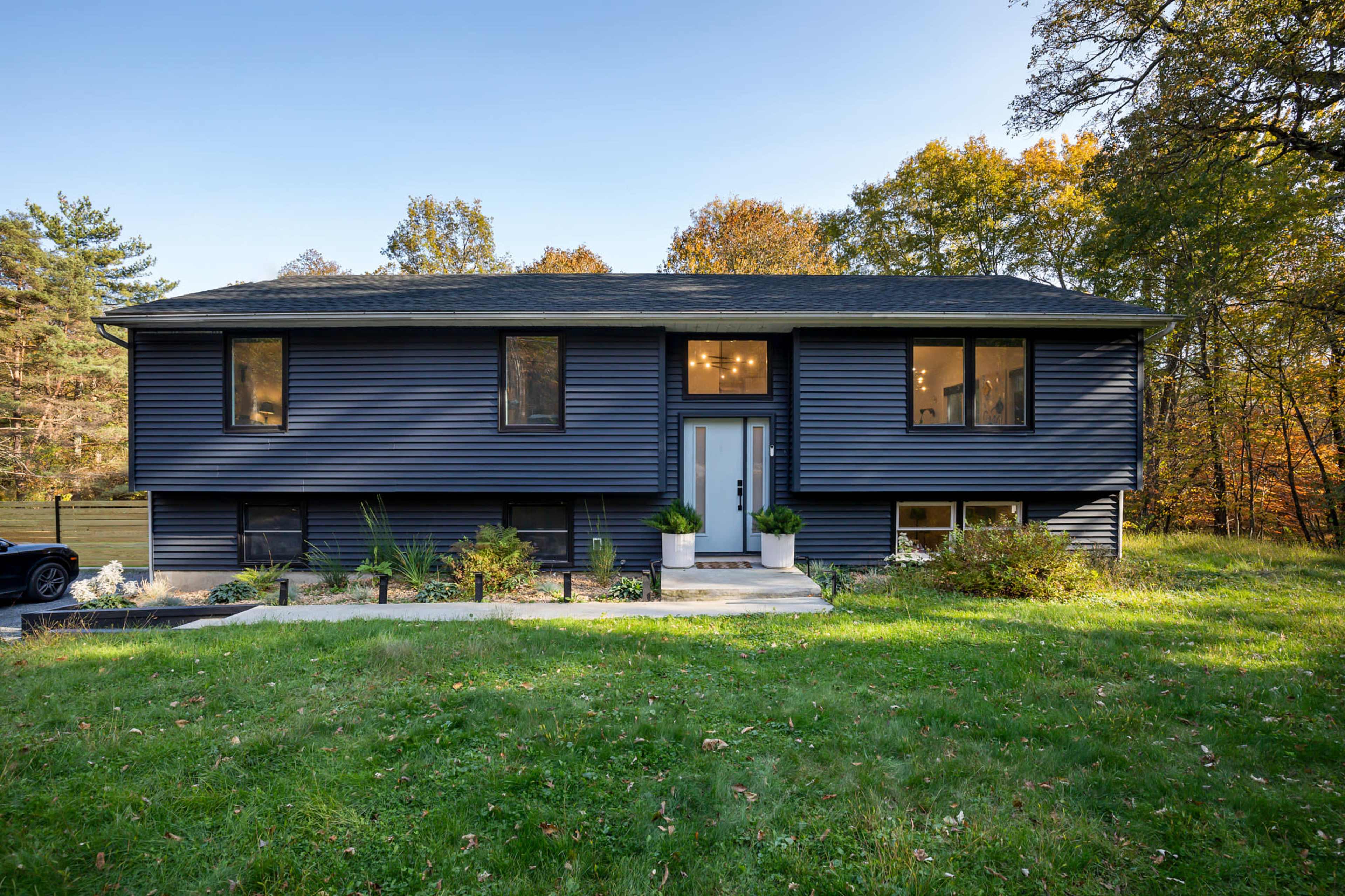 A dark blue, two-story house with large windows and a front porch is situated on a grassy lawn surrounded by trees.