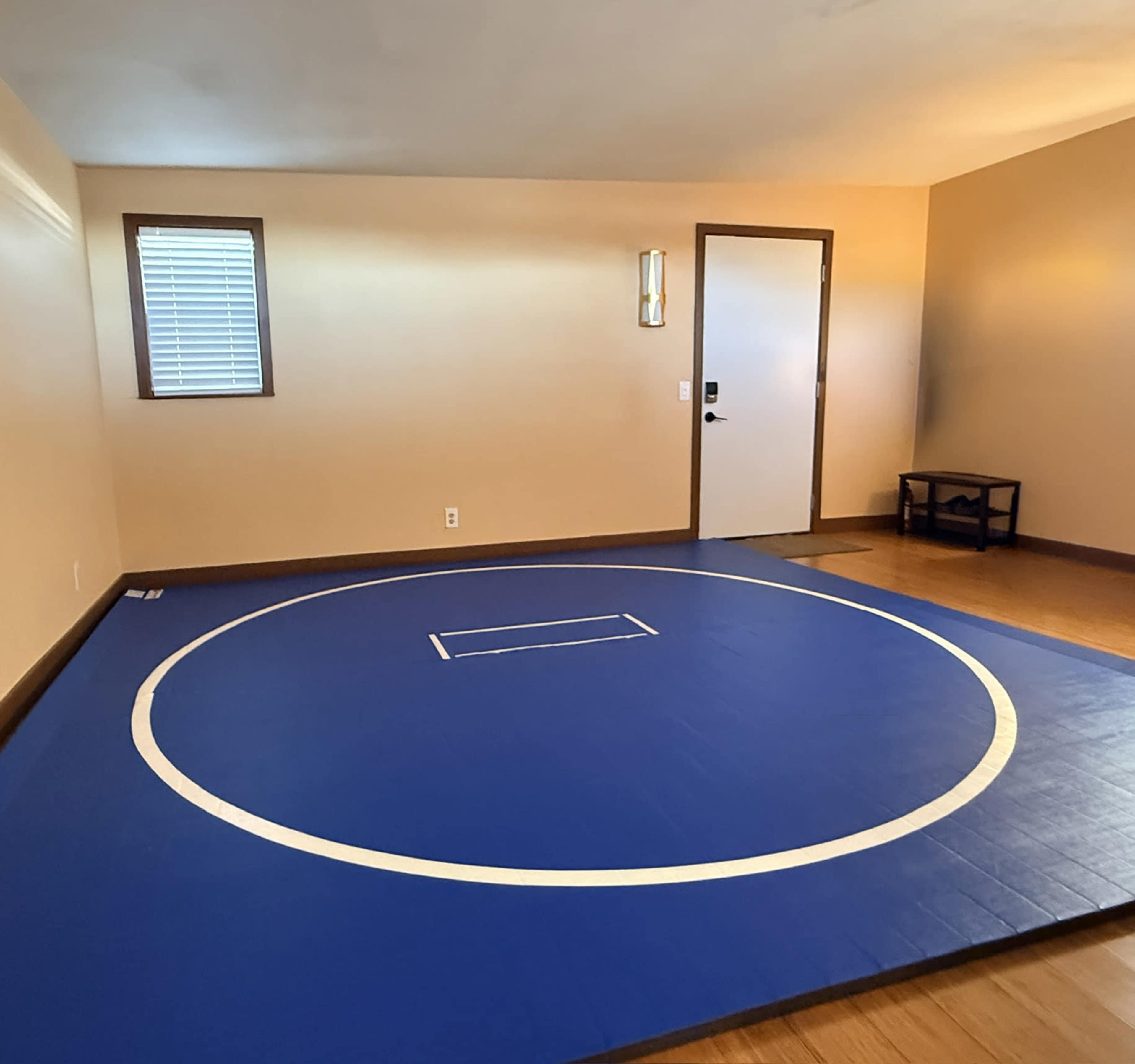 The image shows a room with wooden flooring that features a blue wrestling mat with a white circle and a small rectangle in the center.