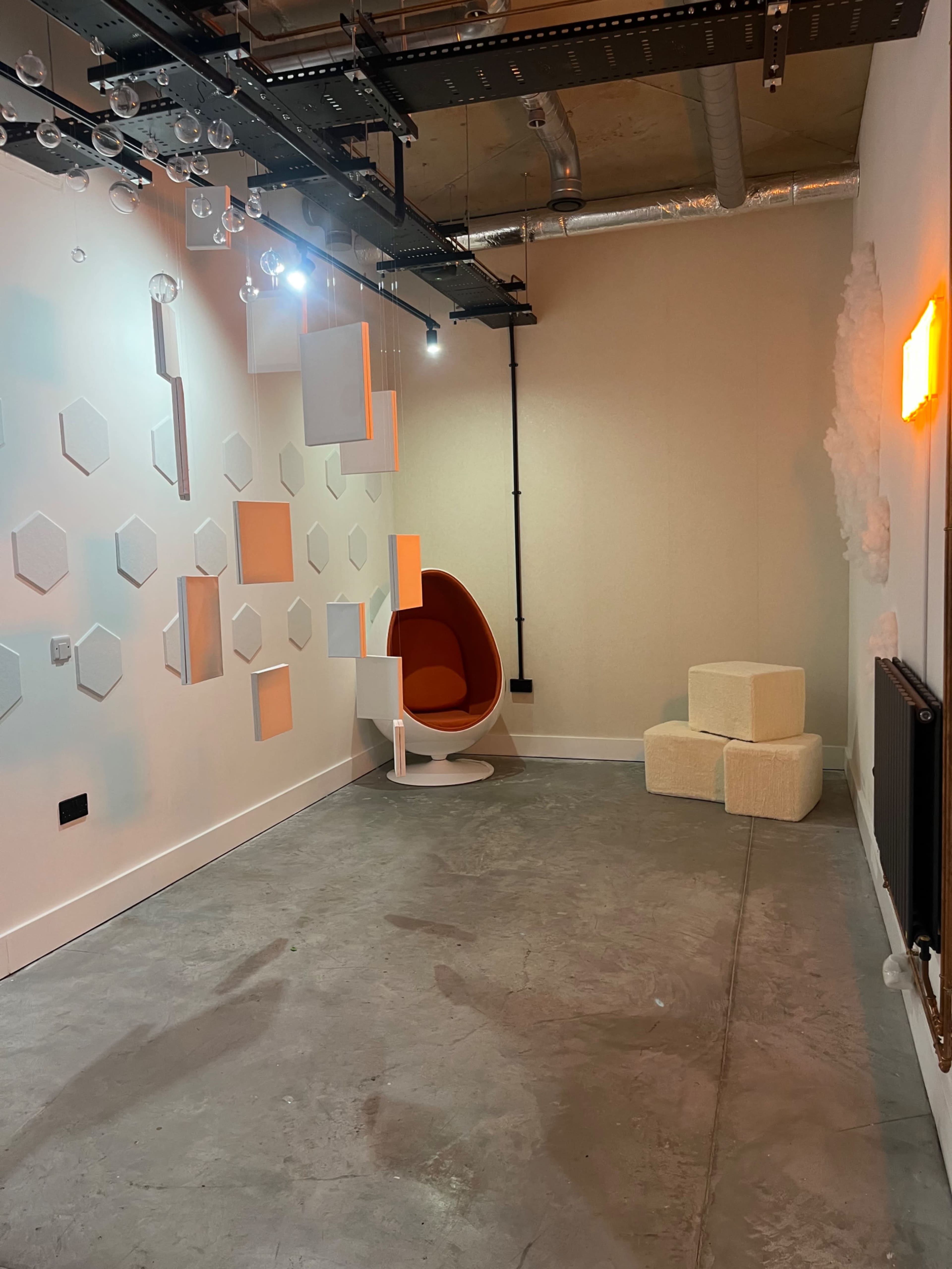 The image shows a minimalist interior space with a red egg-shaped chair, geometric wall panels, and light-colored seating blocks.