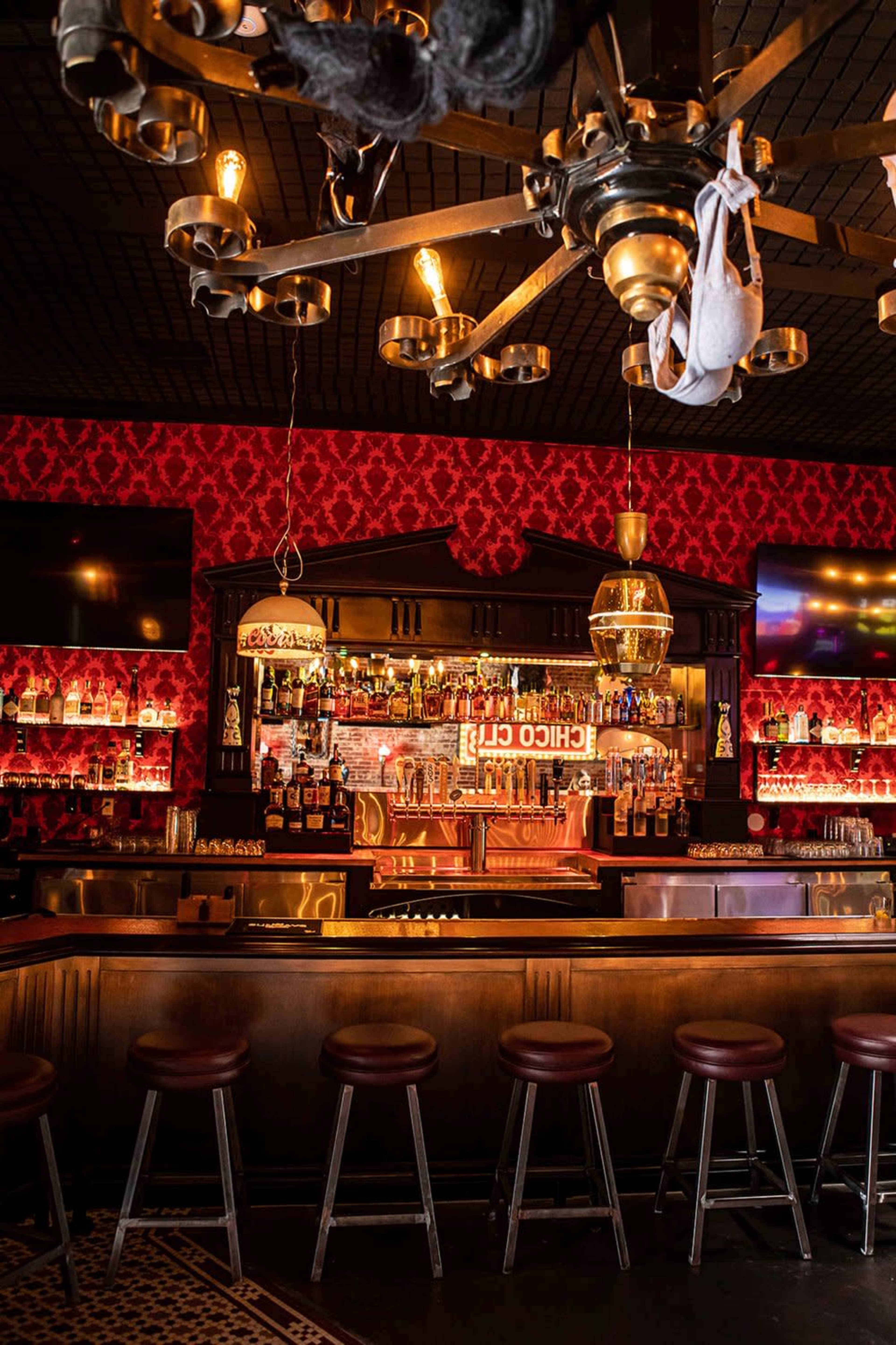 The image shows a bar with a wooden counter, high stools, and illuminated shelves filled with bottles, against a backdrop of red patterned wallpaper and vintage light fixtures.