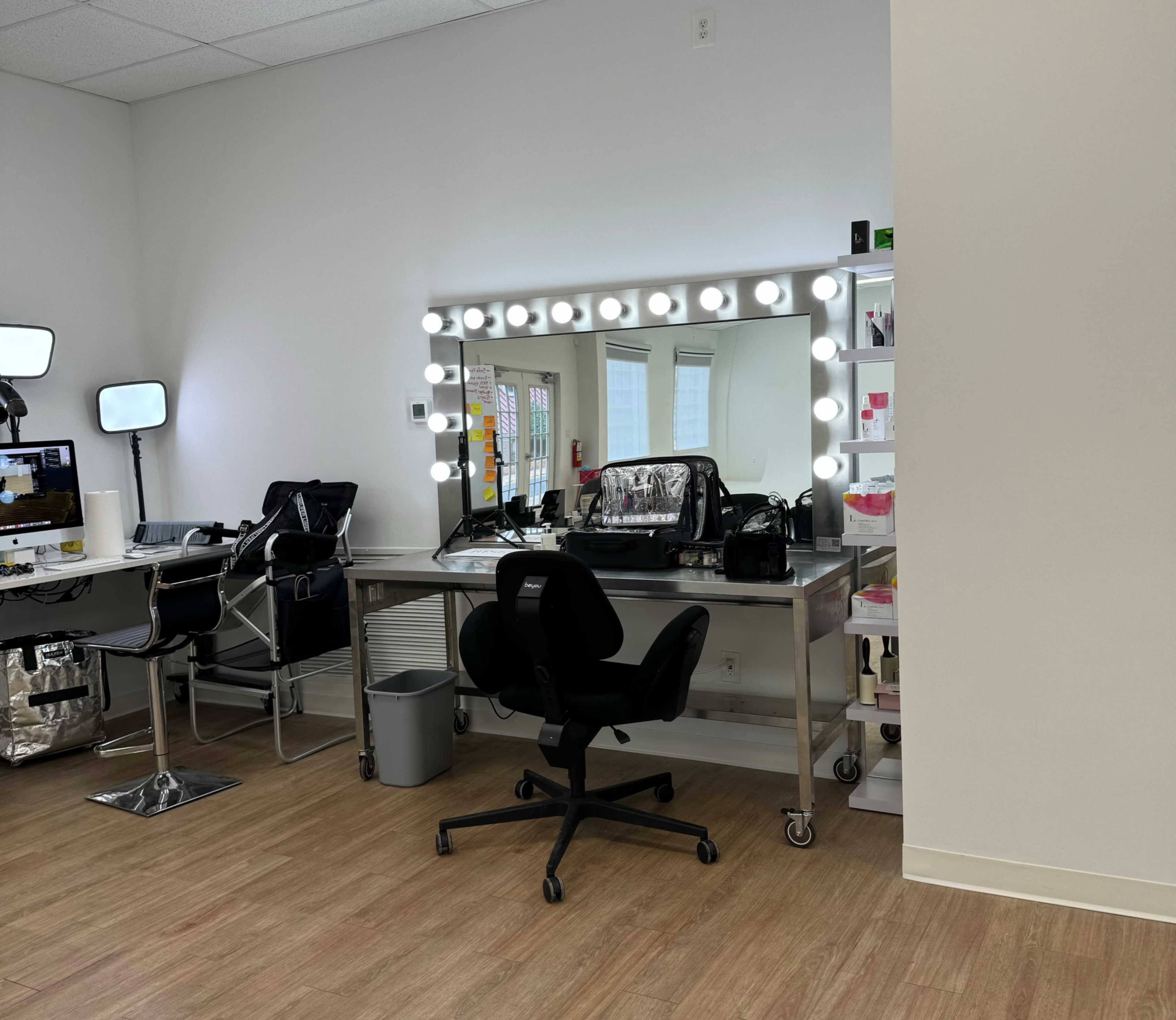 A well-lit makeup station features a large mirror surrounded by lights and various beauty products on a table, with a black rolling chair nearby.