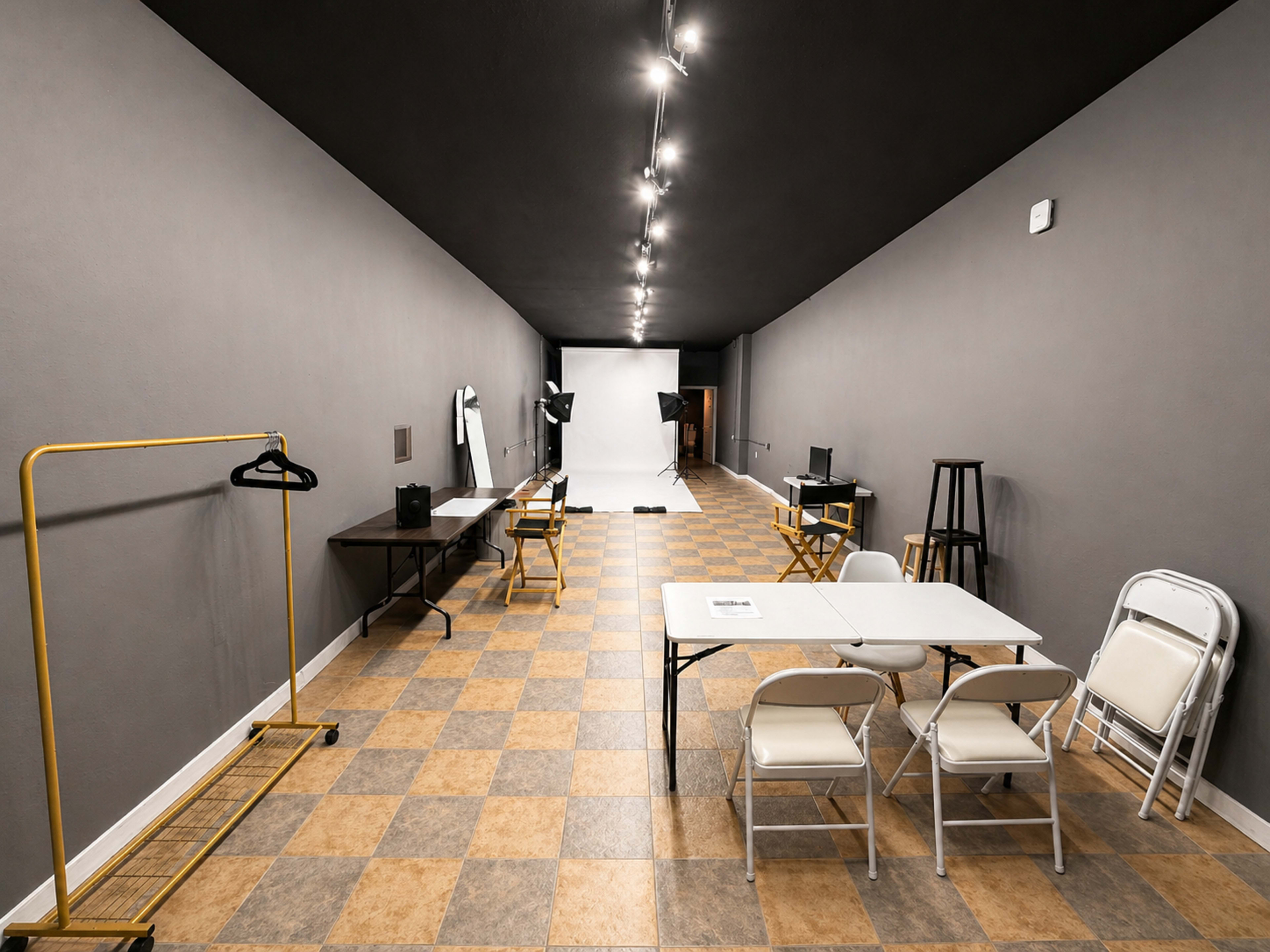 The image shows an empty studio space with gray walls, checkered flooring, and several tables and chairs arranged in a long, narrow layout, with lighting and equipment set up at one end.