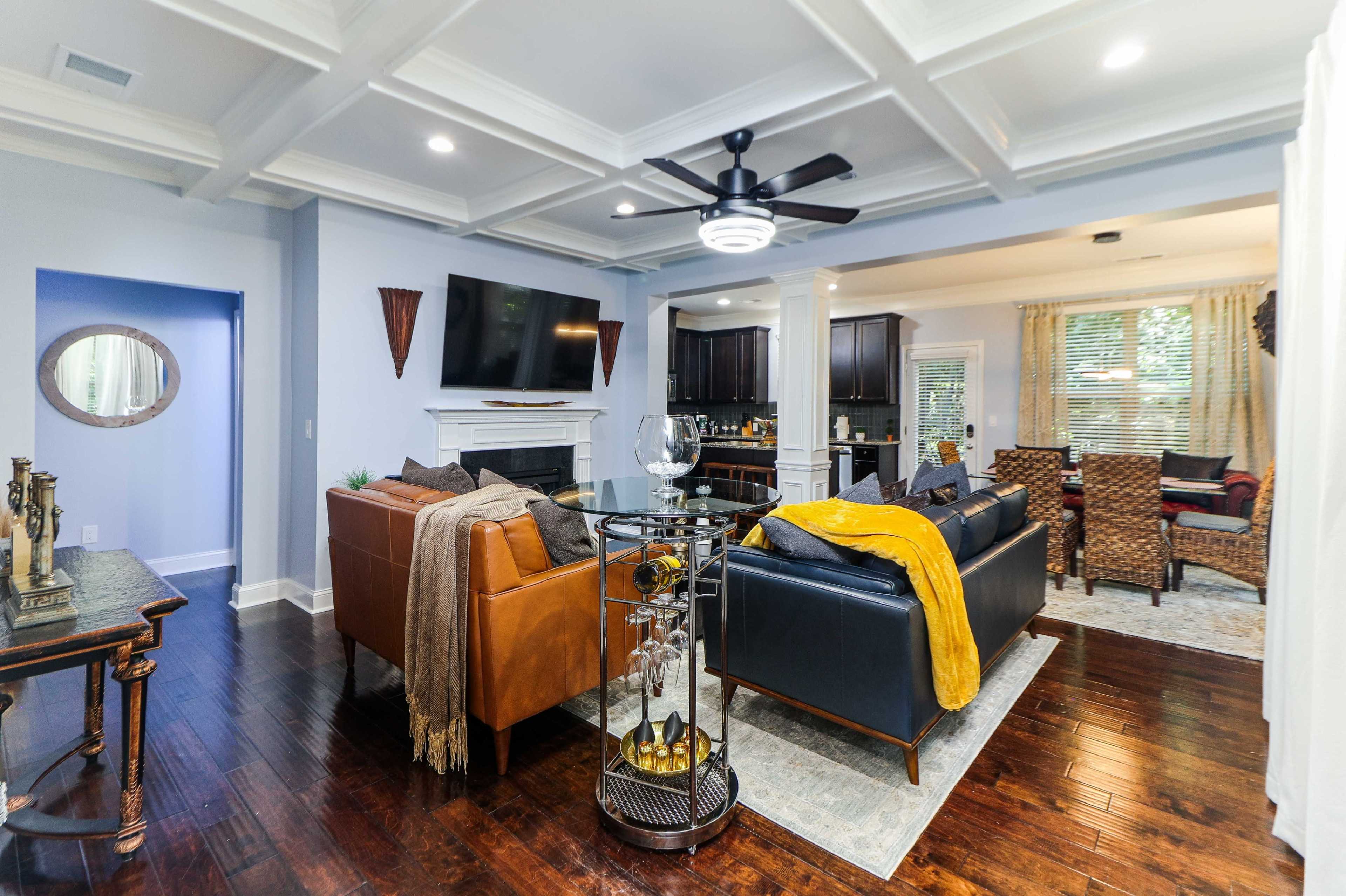 A modern living room with a mix of brown and black sofas, a ceiling fan, a flat-screen TV above a fireplace, and a dining area in the background.