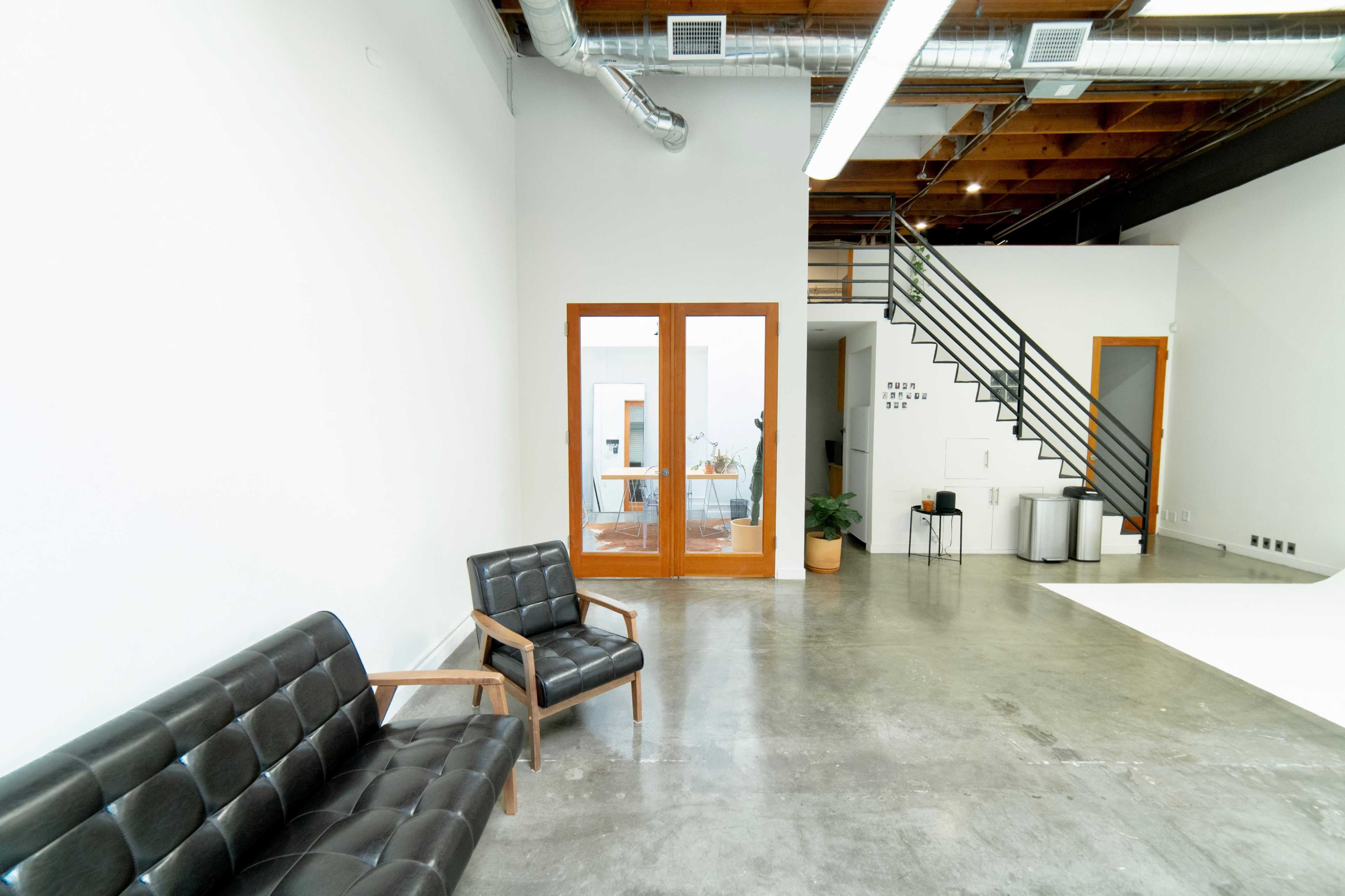 The image shows a modern interior space with a black leather sofa, a wooden armchair, a staircase, and large glass doors leading to another room.